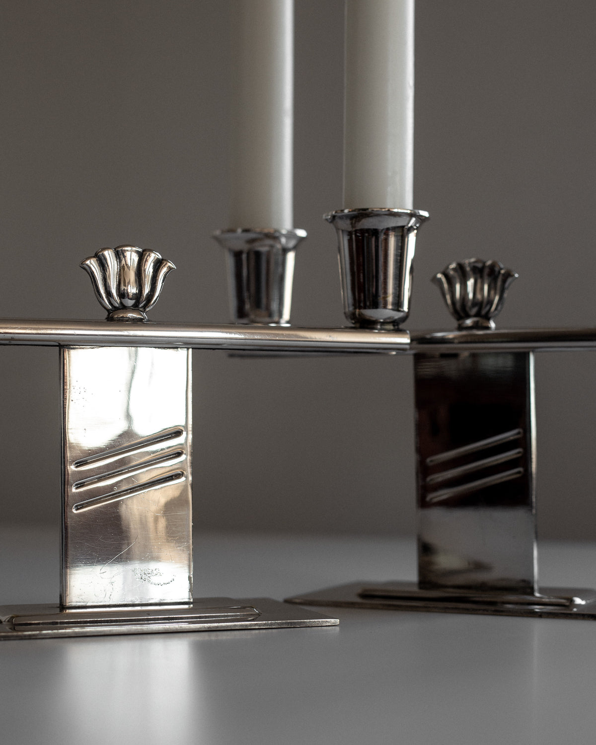 A Pair of Silver-Plated Art Deco Candle Holders from Spigel, featuring a sleek, modern design, stand on a reflective surface. Each holder cradles a white candle with intricate detailing on the candle cups. The softly blurred background emphasizes the shiny metal finish and suggests hints of Scandinavian craftsmanship.