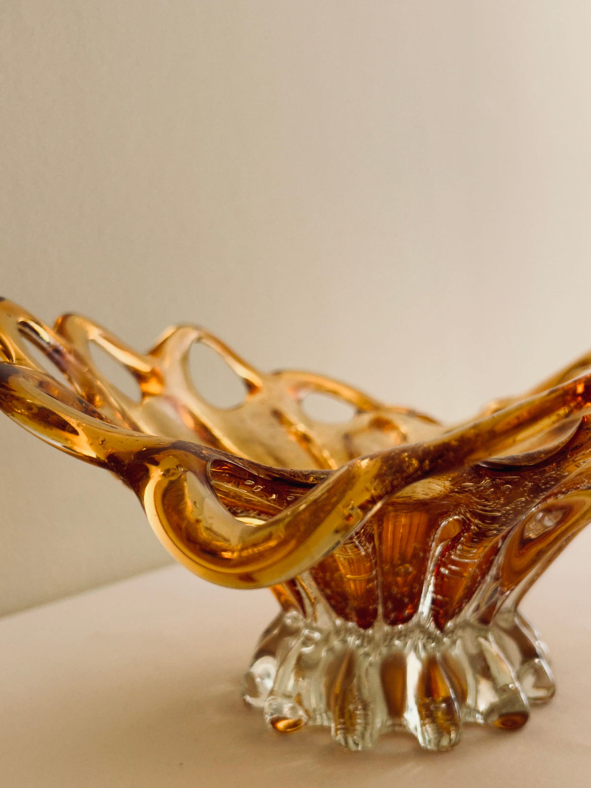 The Murano Glass Amber Bowl by Flock Studio is a masterpiece of intricate Italian glass artistry, featuring an amber and clear design. Its elegantly curved loops and fluted base make a stunning statement against a neutral backdrop.