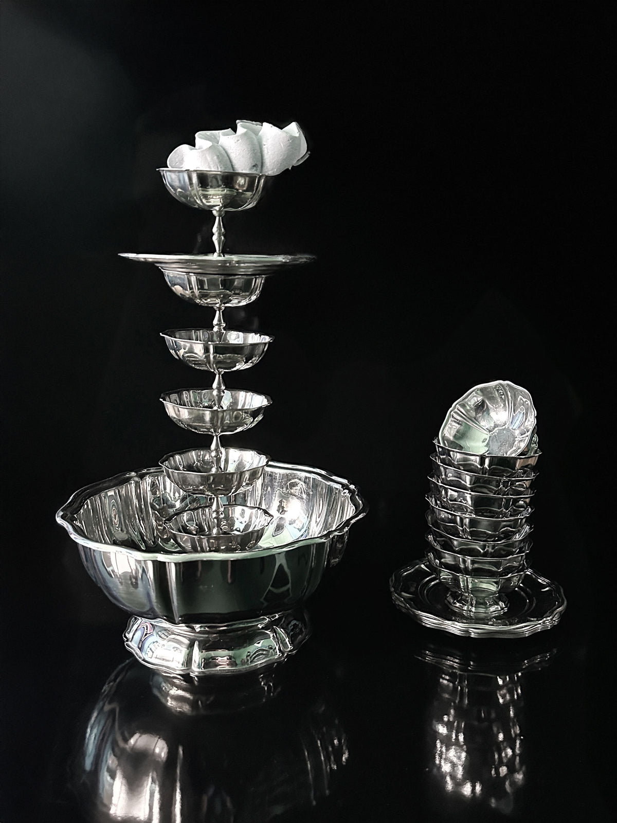 The Dodo Vintage Italian Stainless Steel Dessert Coupe Set features a tiered display of bowls with a single white sugar cube atop. A stack of silver cups enhances the elegant arrangement against a black backdrop.