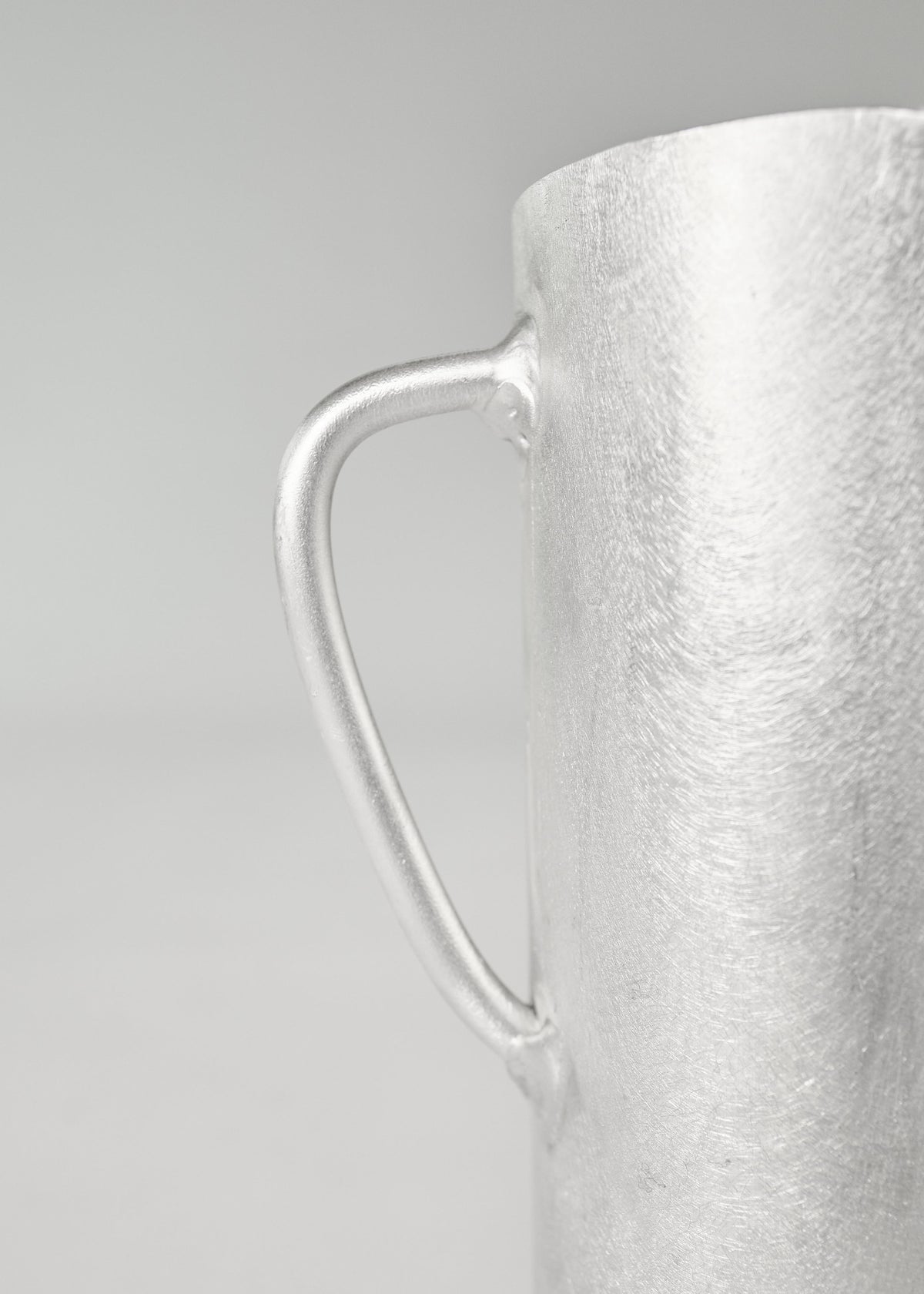 Close-up of Six Dots Design's "The Aluminum Jug," a handcrafted metallic jug with a smooth texture, showcasing its handle on the left side against a plain, light background. This exquisite piece originates from a North London studio, reflecting expert craftsmanship in every detail.