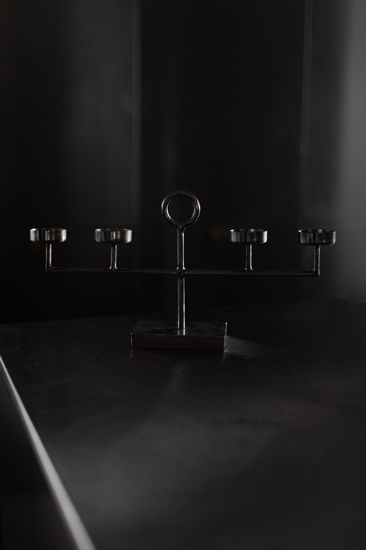 A dark, atmospheric image features the Steel Candleholder by Out For Lunch, showcasing its five elegantly curved arms and candleholders on a reflective surface. The dimly lit background adds a moody, modern minimalistic vibe.