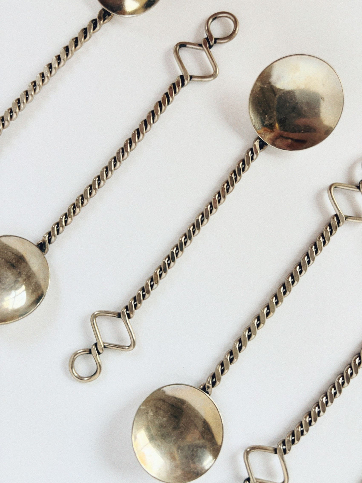 Close-up of Huerto's 70's Handmade Spoons Set with twisted handles and looped ends, displayed diagonally on white. These spoons showcase a reflective metallic surface and unique geometric designs, capturing the essence of functional art.
