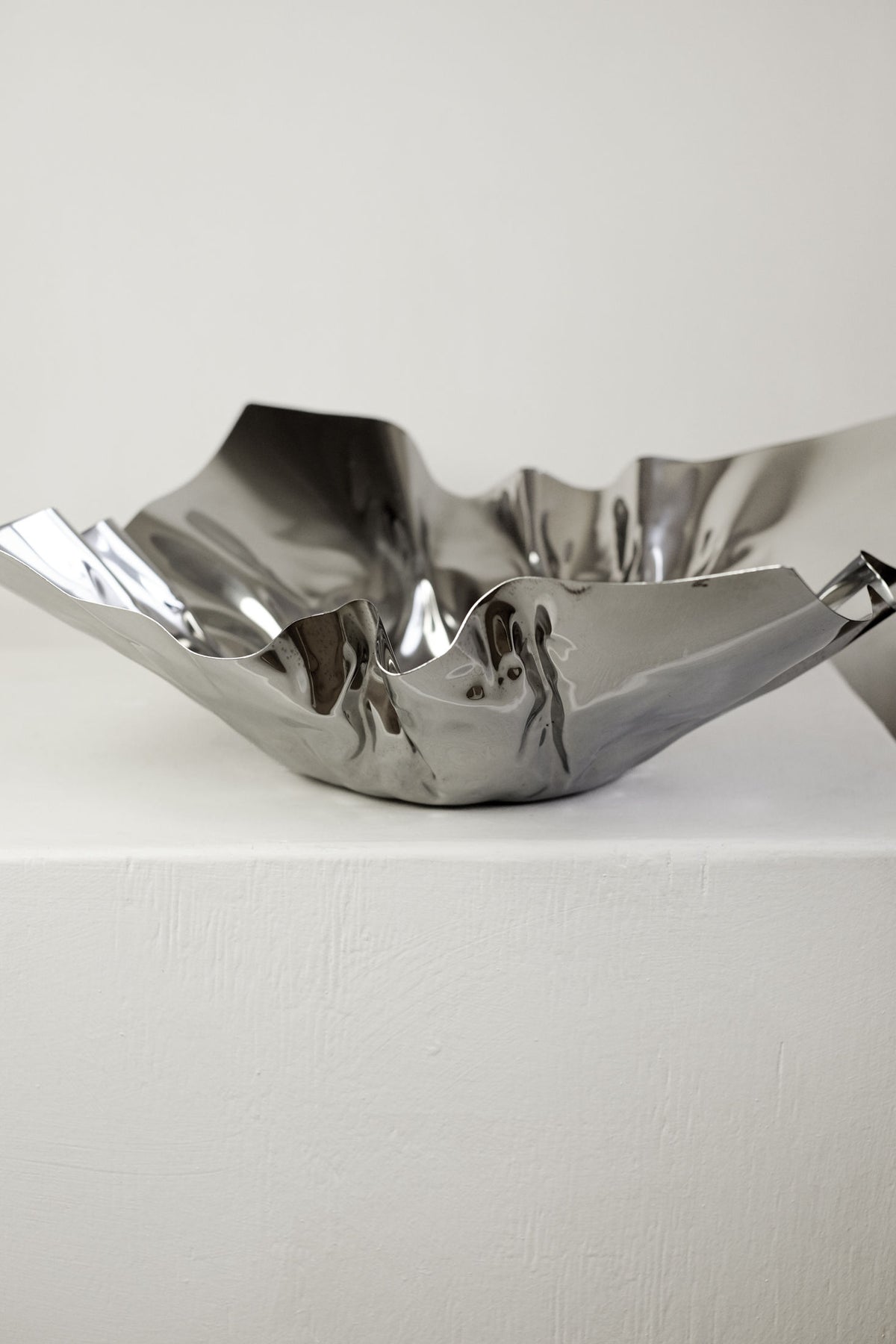 The Extra Large Folded Chrome Bowl by septembre studios, featuring a shiny, crumpled finish and wavy edge, adds vintage charm to any room as it sits on a white textured surface against a plain light background.