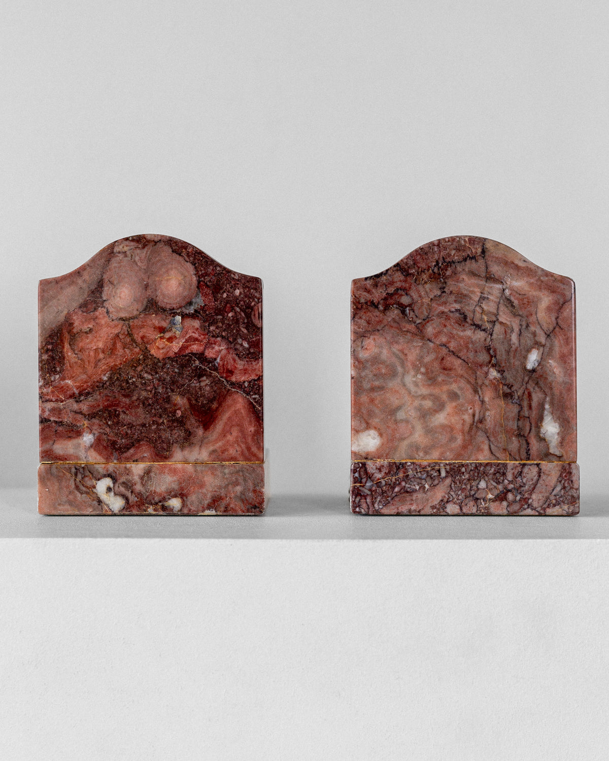 Spigel's Pair of Art Deco Marble Bookends, crafted in reddish-brown marble with intricate natural patterns, stand on a white surface. Their arched tops display unique earthy swirls and veins, adding geometric flair against a plain gray wall.