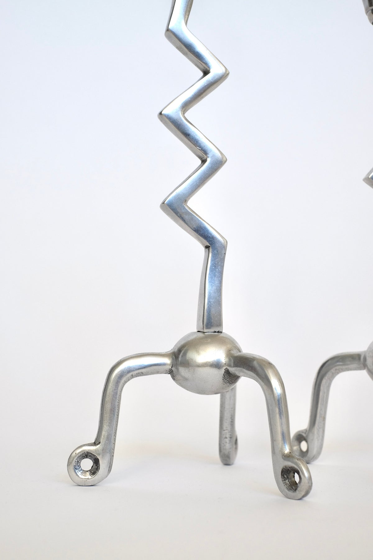 A close-up of the "Set of Zigzag Candleholders" by septembre studios showcases a silver metal sculpture, reminiscent of rare find candle holders. It features a zigzag shape extending upward, connected to three curved legs with small holes at their ends. The minimalist and abstract design is set against a plain white background.