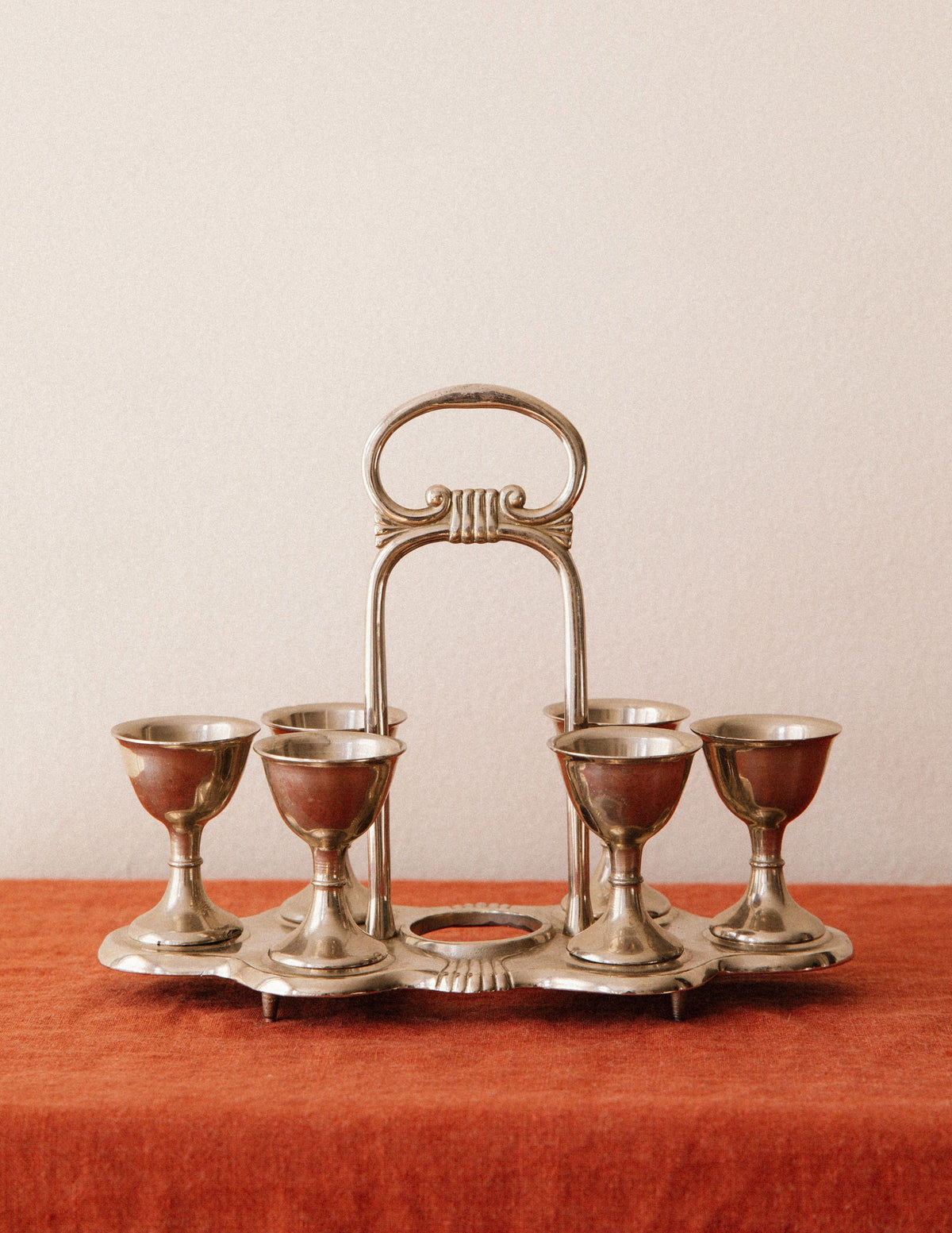 The Arcā Collections Vintage Egg Cruet Holder Set features six sculpted silver-plated cups in a circular display, elegantly presented on a rust-colored cloth before a plain beige wall.