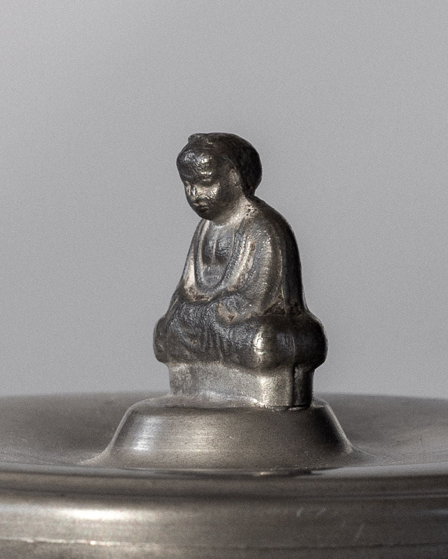 A close-up of a small, metallic Buddha figure sitting cross-legged atop a rounded surface, reminiscent of the Spigel Art Deco Metal Box from 1934, set against a plain background. The intricate detailing on its robe and serene facial expression reflect the elegance associated with Svenskt Tenn designs.