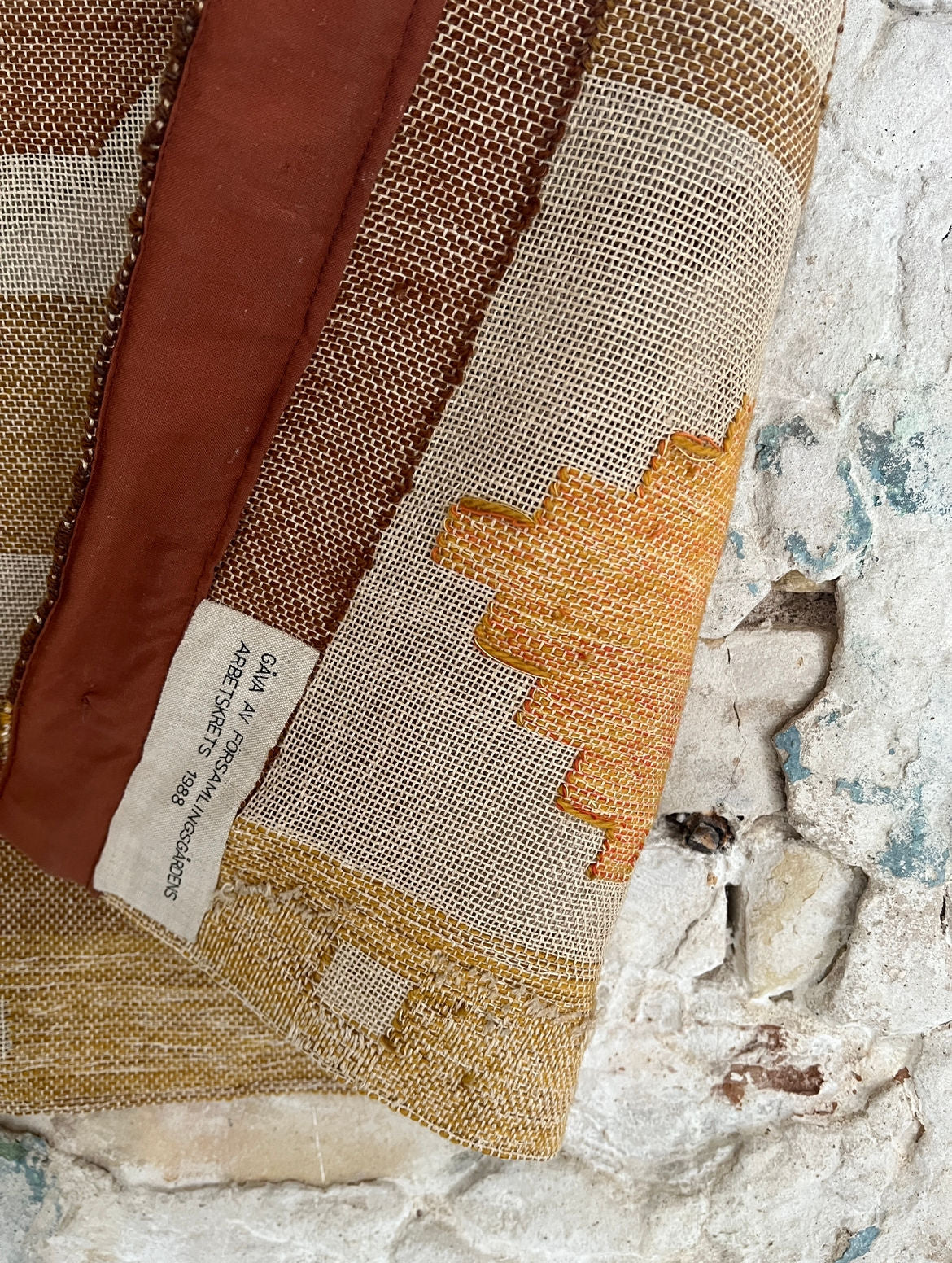 A close-up of the Large Abstract Wool Wall Tapestry by Veter Vintage (Sweden, 1988) showcases brown and golden yellow geometric patterns with a brown border, plus a fabric care tag, draped over textured weathered stone.