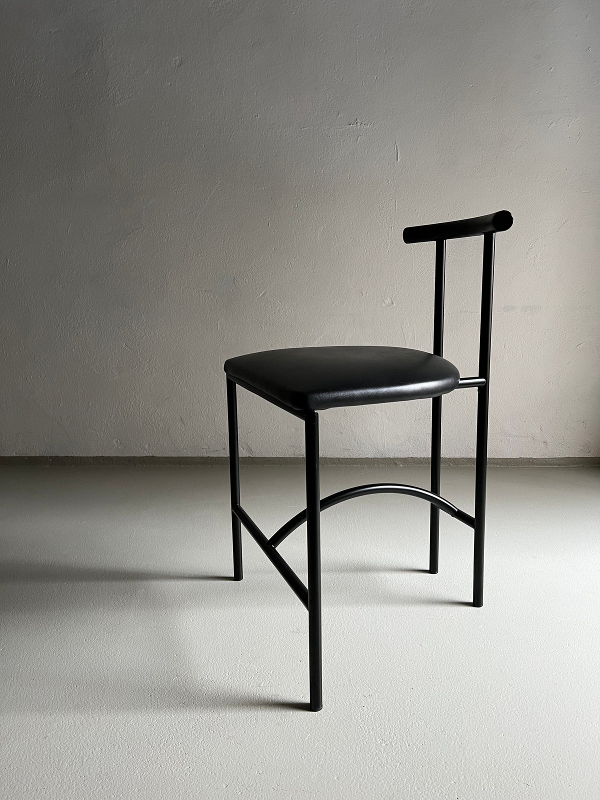 A black metal chair with a minimalist design stands against a plain, light-colored wall. The vintage-condition 4 Postmodern "Tokyo" Chair by Rodney Kinsman | Italy | 1980s from Veter Vintage features a padded black faux leather seat and a simple T-shaped backrest, casting a subtle shadow on the smooth, neutral floor.