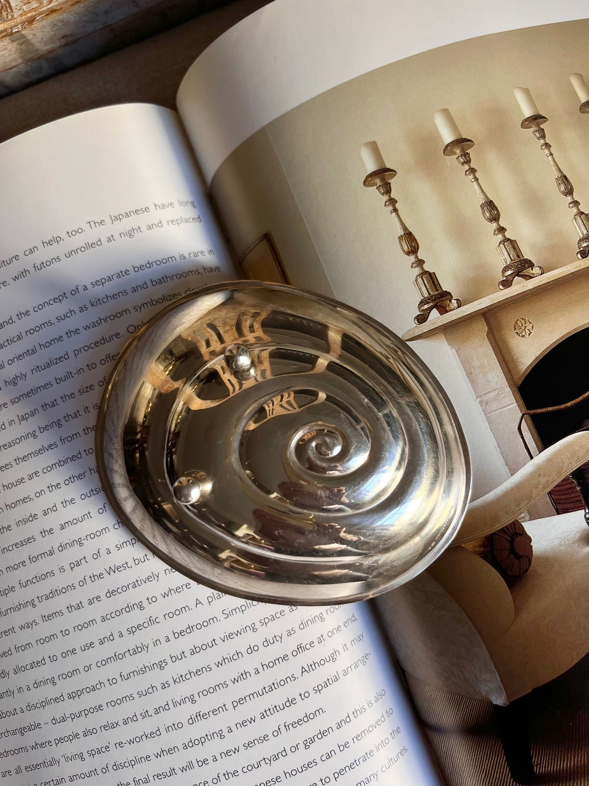 A rare sterling silver Midcentury Vide-Poches by Médecine rests on an open book. The book displays text on the left page and a cozy living room scene on the right, featuring sconces and a fireplace. Sunlight illuminates the vide-poches and parts of the book, casting subtle shadows.