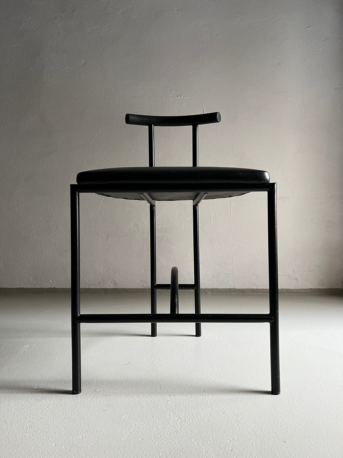 A minimalist black chair with a sleek metal frame and a low backrest, upholstered in faux leather, is positioned against a plain, light gray wall on a smooth, light-colored floor. The 4 Postmodern "Tokyo" Chairs by Rodney Kinsman | Italy | 1980s from Veter Vintage feature clean lines and a modern aesthetic.