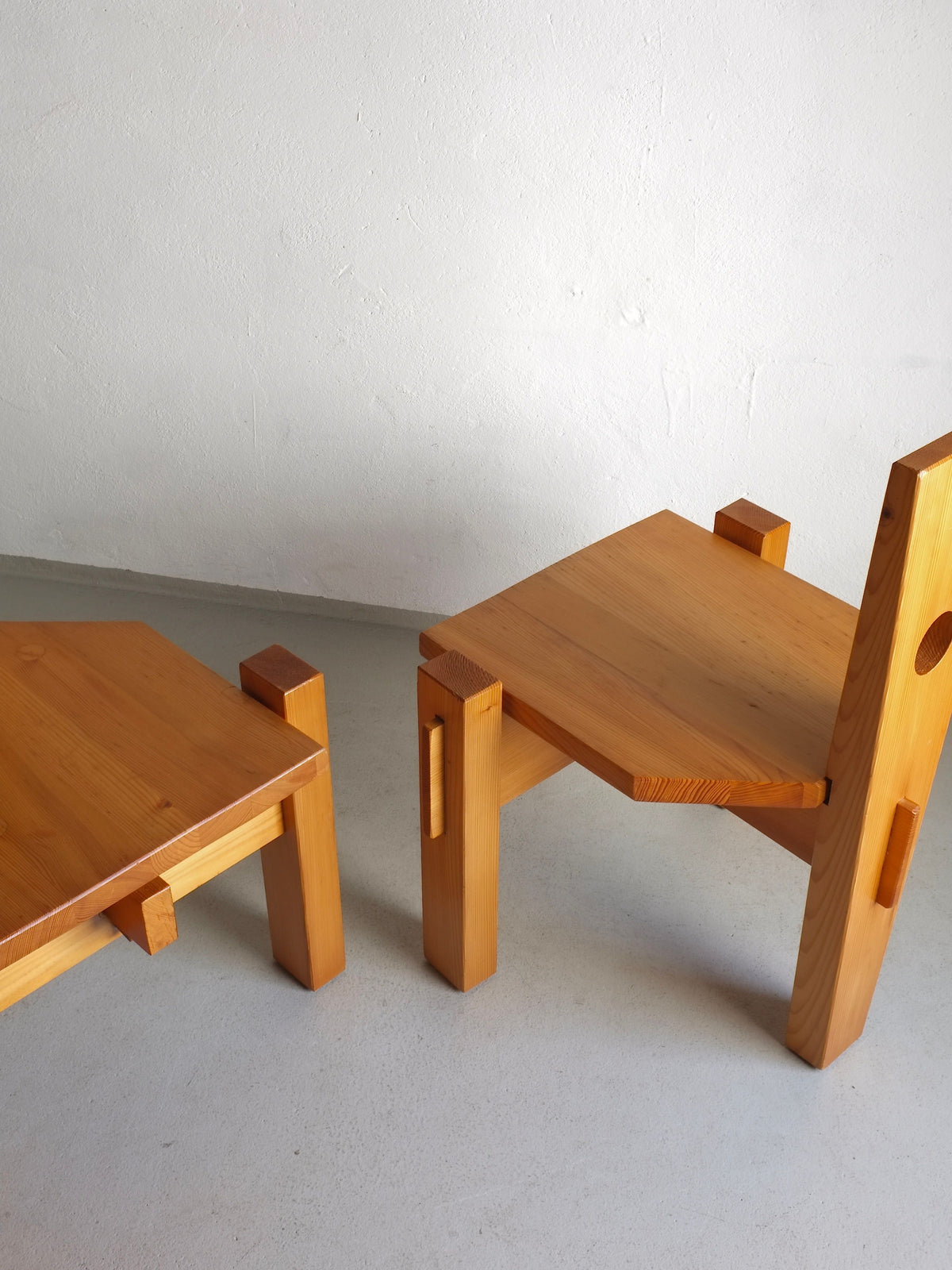 Two Veter Vintage 1960s sculptural pine chairs by Erwin Egel, one with a tall backrest and the other a stool, feature a minimalist geometric design with thick rectangular legs. They are placed side by side on a light gray floor against a white wall.