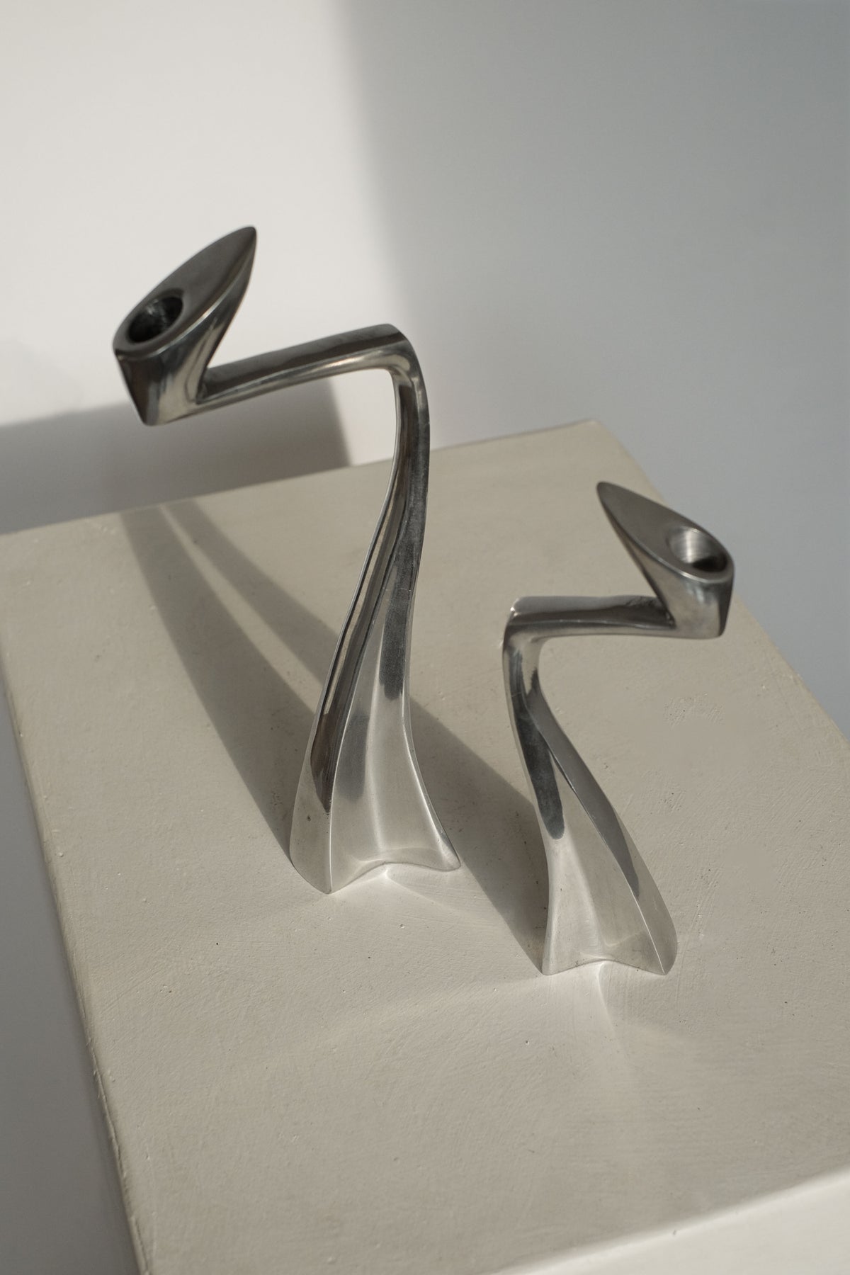 A pair of sleek, modern candleholders by Matthew Hilton rests on a light surface, their curved designs and shiny finish highlighted by soft natural light. Perfect for design enthusiasts, these pieces from septembre studios evoke elegant 80s sophistication.
