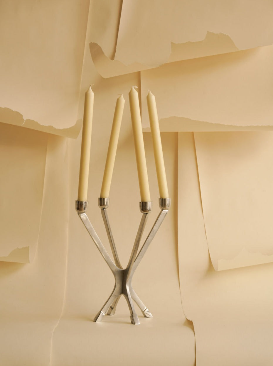 A vintage, silver Les Objets Amsterdam two-piece candelabra with three arms, each holding an unlit beige candle, stands against a textured beige paper background with torn layers.