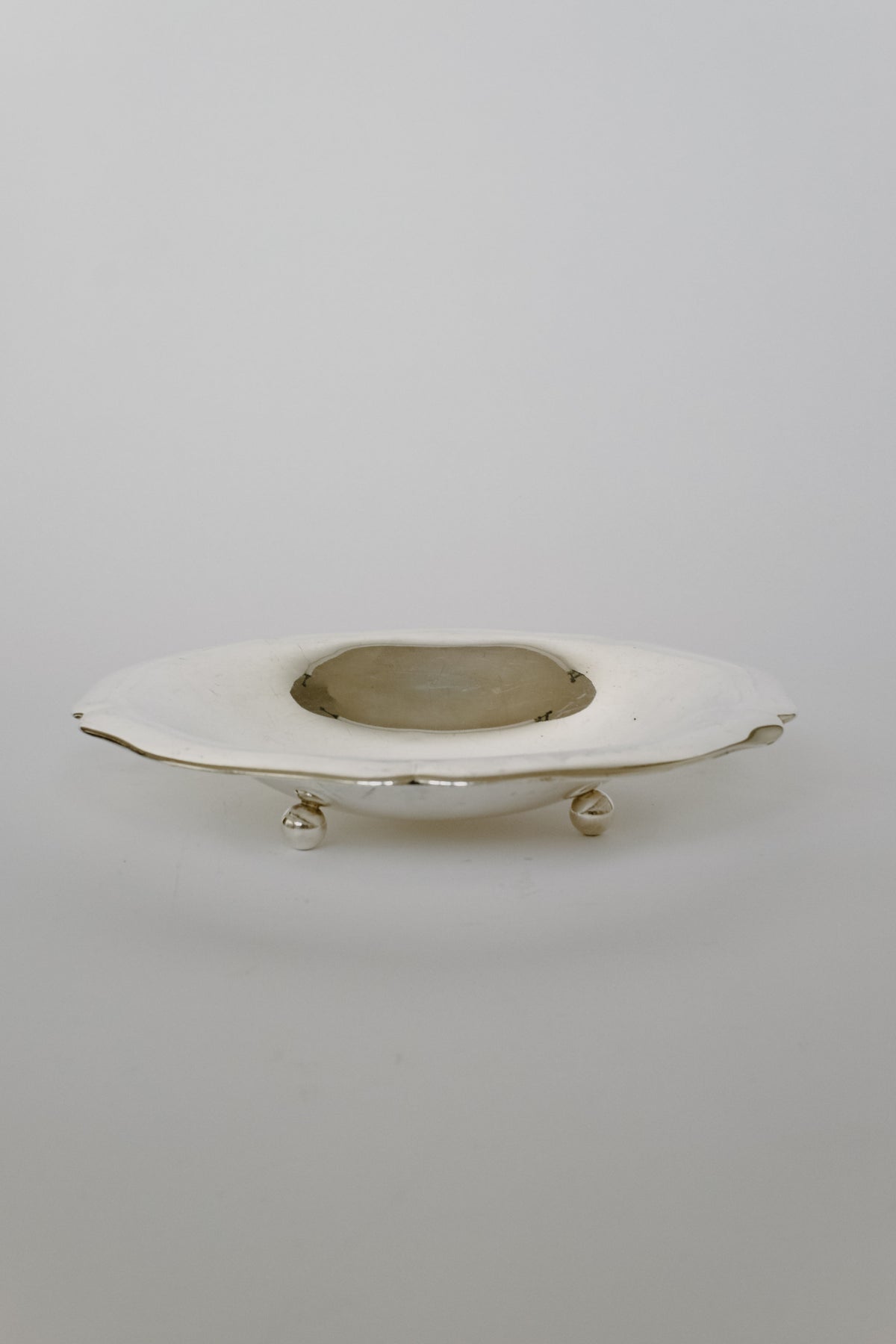 An elegant, oval-shaped Silverplated Bowl from septembre studios with a vintage charm and intricate detailing is placed against a simple background. The bowl is elevated on small decorative feet, and its gleaming surface reflects the light softly, bringing distinctive character to any environment.
