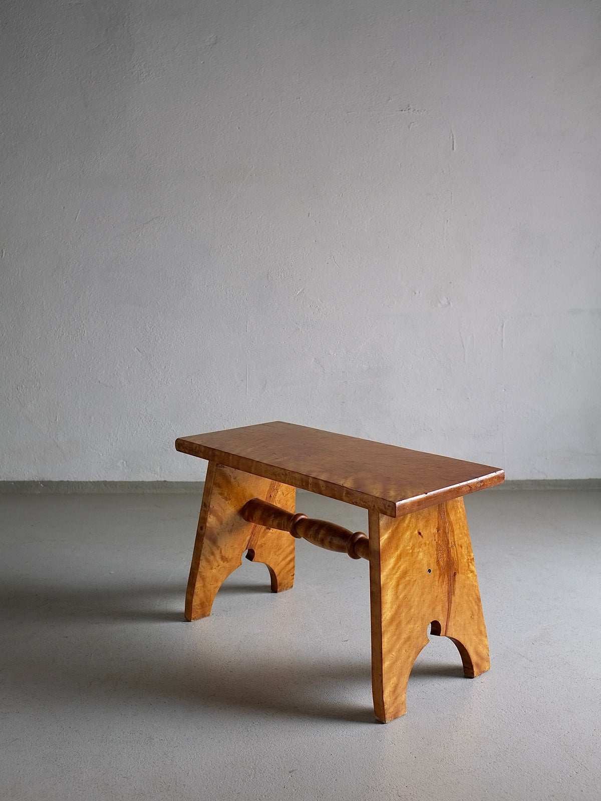 A small wooden stool carved from solid Karelian birch wood with a polished finish featuring a simple design, a flat rectangular seat, and two curved legs connected by a horizontal spindle. The Rustic Solid Karelian Birch Stool | Latvia | 1950s by Veter Vintage is placed on a smooth, light gray floor against a plain, light-colored wall.