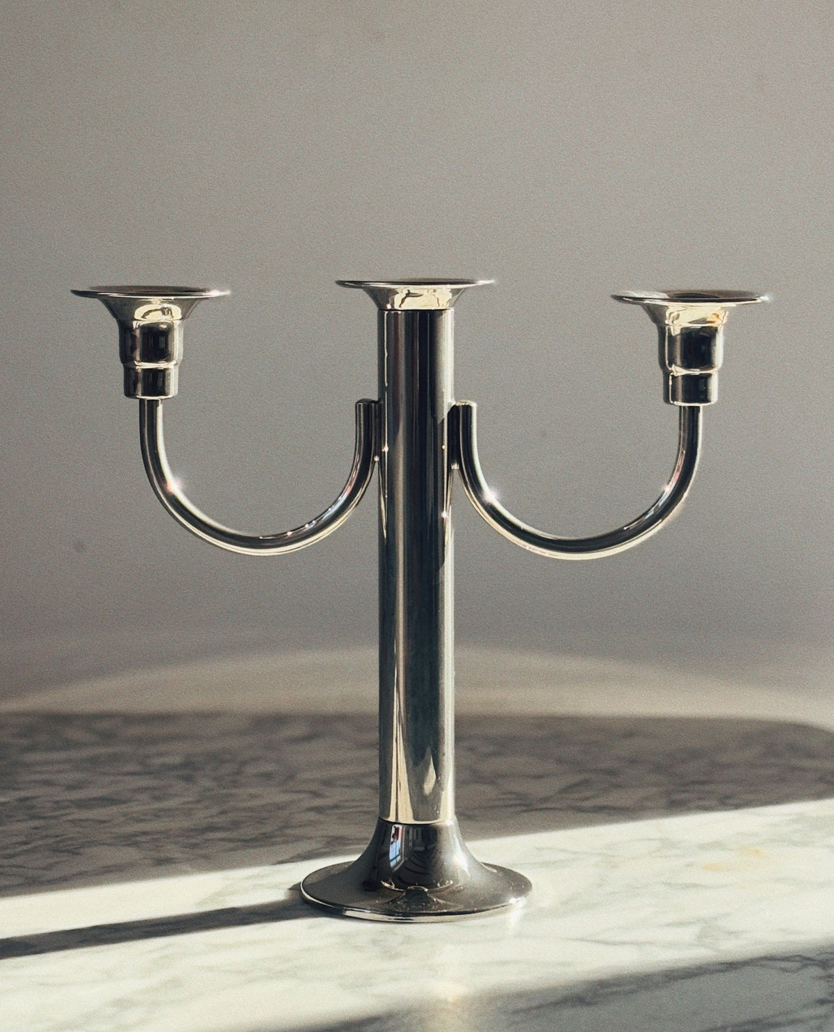The Huerto 3 Arm Metal Candleholder, finished in silver, stands elegantly on a marble surface casting soft shadows against a plain neutral wall.