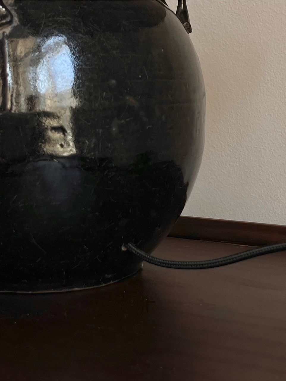 The Médecine Antique Glazed Lamp with Ears, a sleek black ceramic piece reminiscent of an antique vessel, sits on a dark wooden surface. A thin black cord extends from its bottom right. The background features a plain white wall.