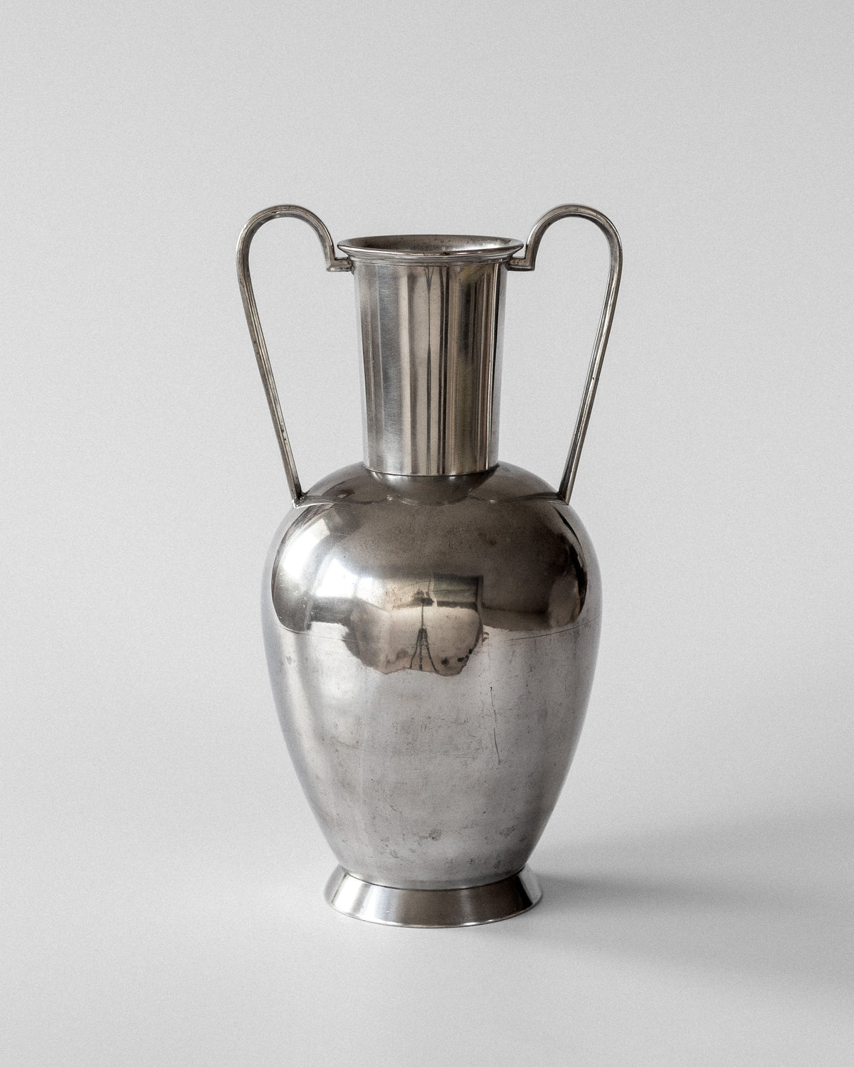 The Large Modernist Pewter Amphora by Spigel, from the 1930s, features tall curved handles and a reflective surface. Its amphora shape has a wide body tapering gracefully to a narrow base and neck, reminiscent of Rolf Engströmer's Scandinavian metalwork.