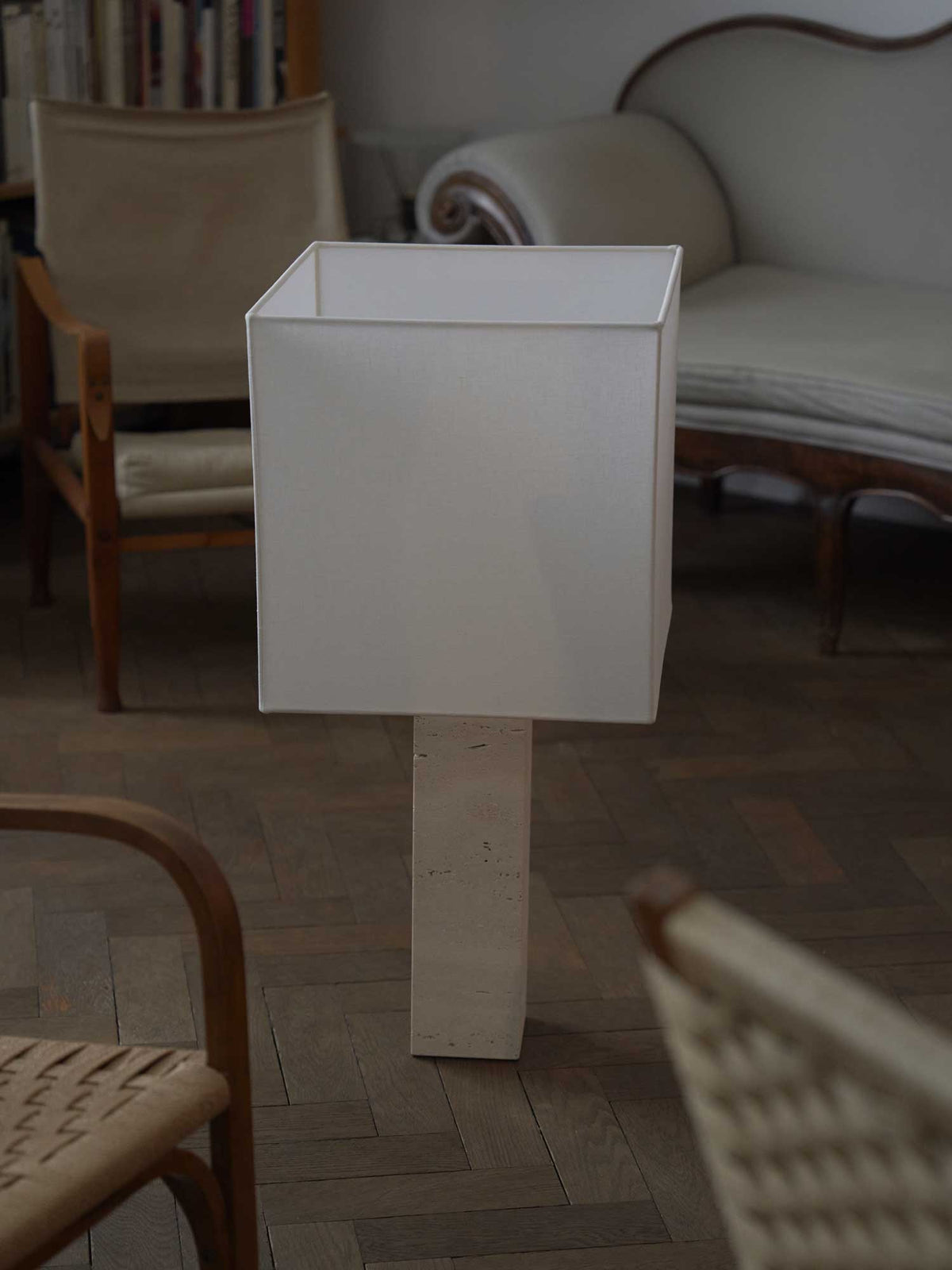 A modern floor lamp, the Roma I Squared White Travertine by Nassi Lamps, with a rectangular white shade and a textured, light-colored base resembling white travertine, is placed on a wooden floor in a cozy, vintage-style room. The room features a wooden chair with a cushioned seat and bookshelves in the background.