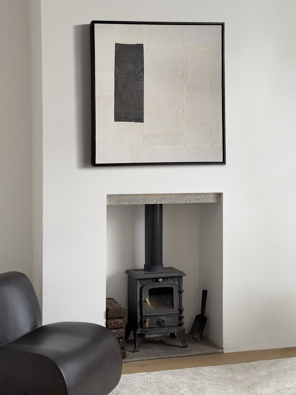A minimalist living room with a black wood-burning stove set into a nook recessed into a white wall. Above the stove is Bec Kirby's (a-tent), 2024 Framed Hand Loomed Tapestry with black and white shapes. A black leather chair is positioned to the left on a light-colored carpet.
