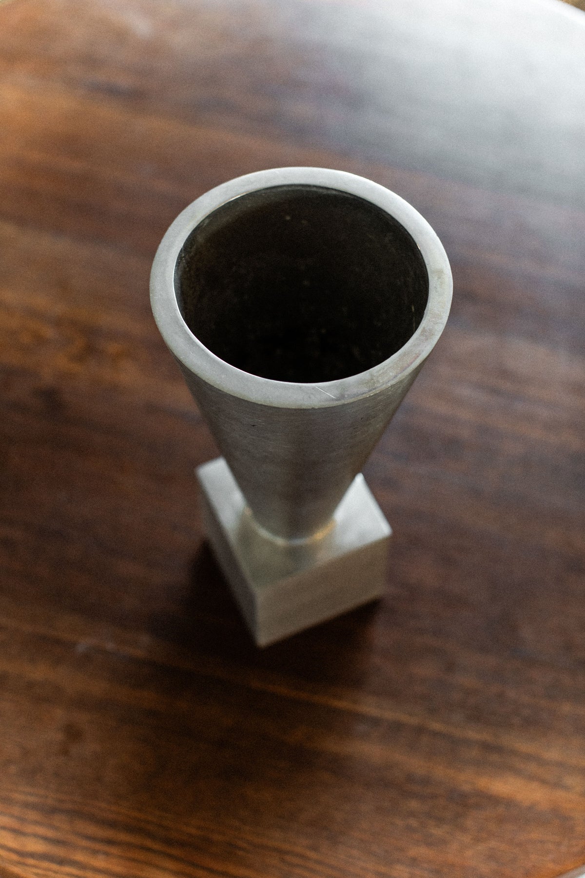 The 70's Aluminum Cast Vase by Out For Lunch, featuring a metallic, cone-shaped design and a square base, sits on a dark wooden surface, viewed from above.