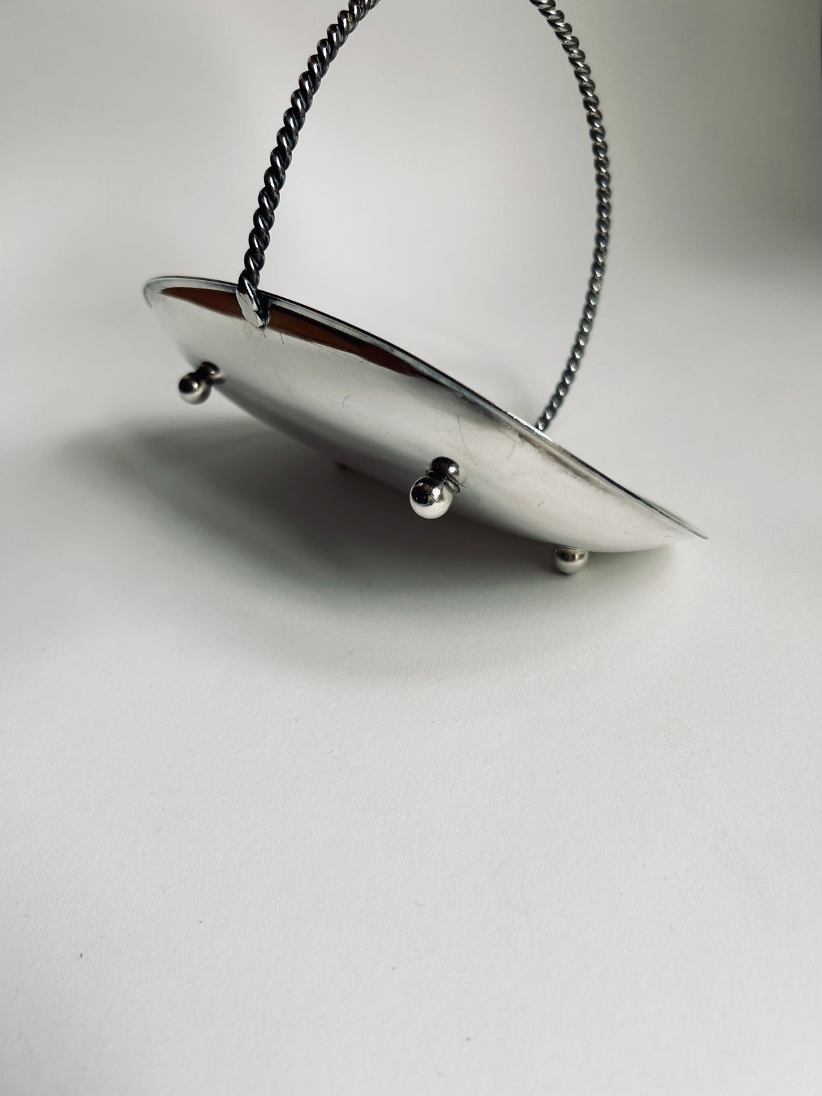 The Art Deco Silver Plated Basket by Flock Studio, featuring a vintage oval design with a twisted handle and four small spherical feet, is set against a plain white background. Its simple yet elegant design exudes timeless sophistication, reflecting light gently across its shiny silver surface.