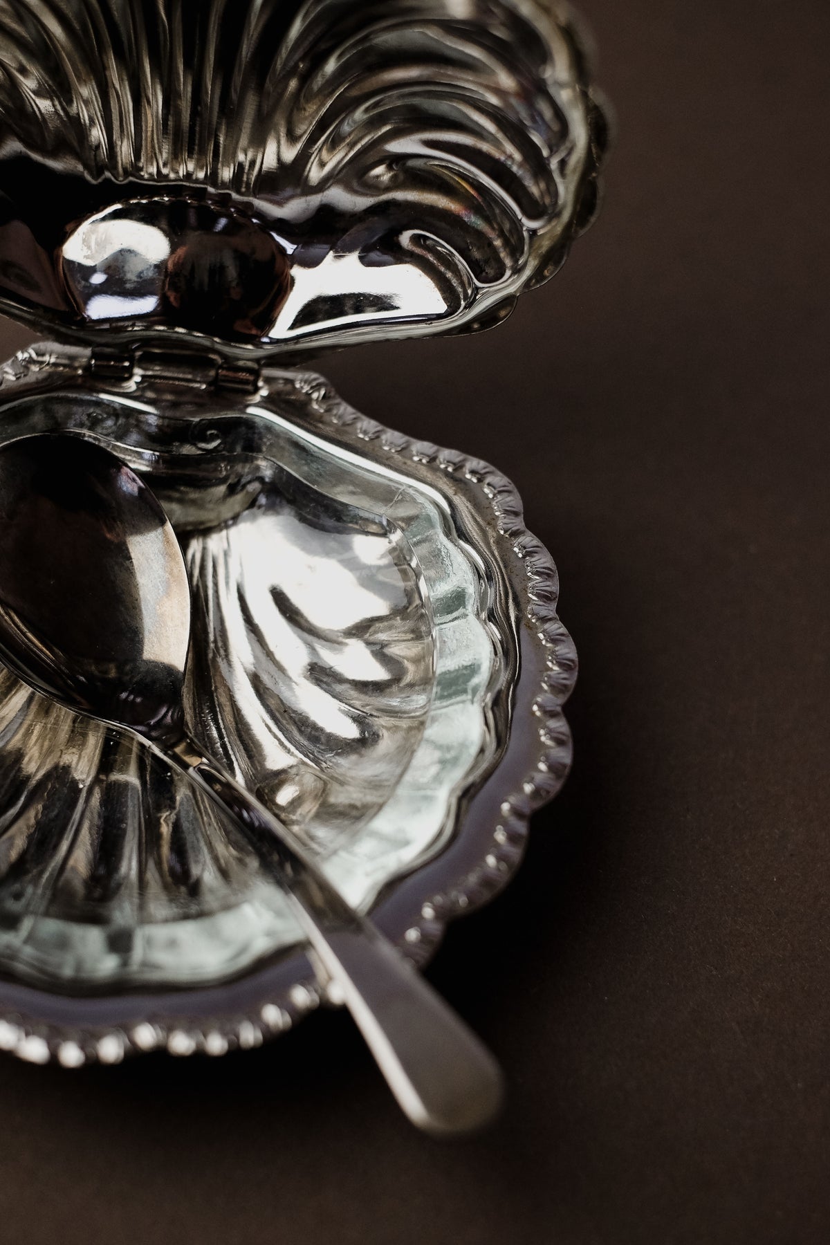 The Shell Shaped Butter Dish with Spoon by septembre studios features a vintage brass design, opening to reveal a shiny interior and an elegantly placed metal spoon. The dark background enhances the reflective surfaces of this exquisite dining piece.