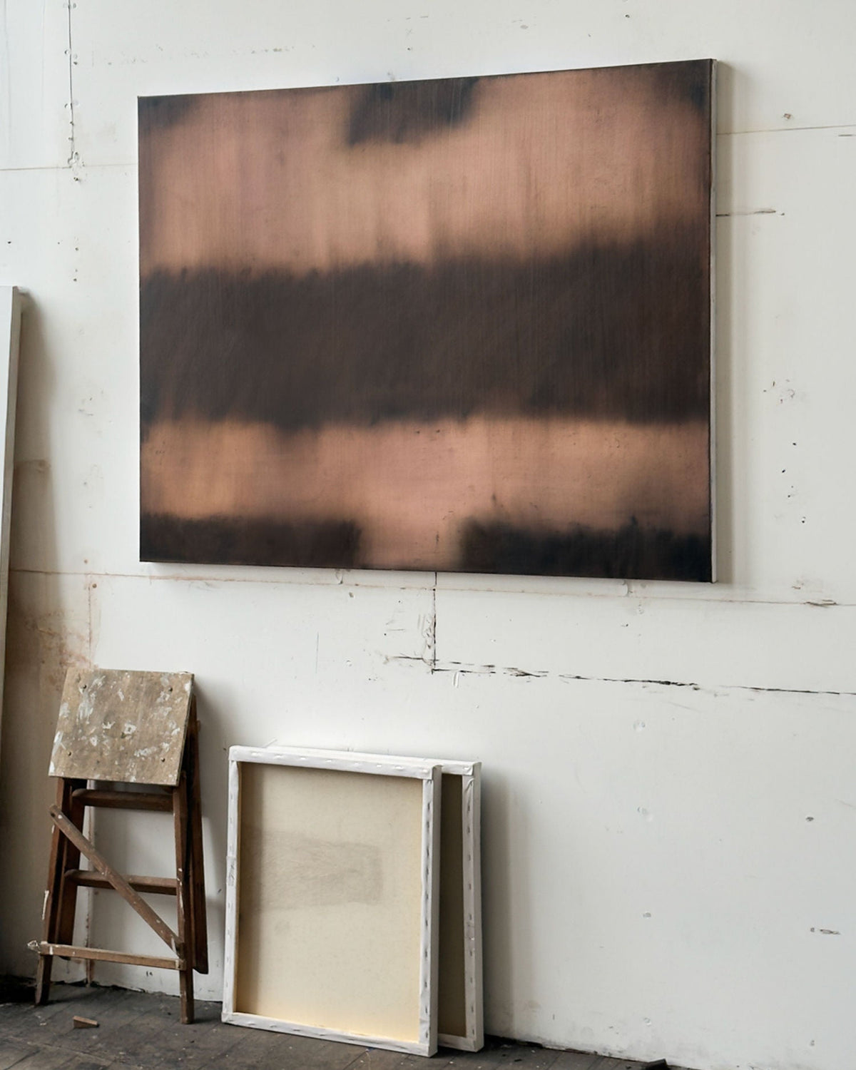 Fever (ST446), 2025 Oil Painting by Bec Kirby, featuring dark and coppery horizontal bands, hangs on a white wall above two empty frames and a worn wooden stool on a rustic wooden floor.