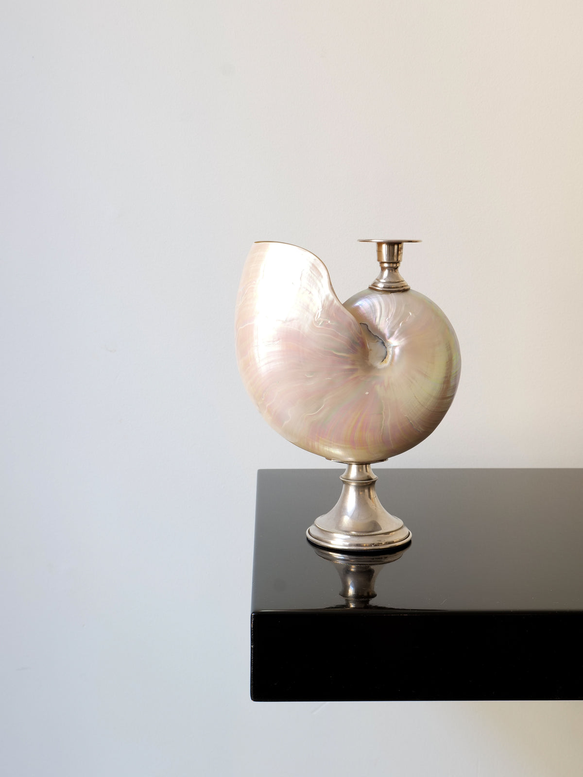 A vase shaped like a nautilus shell, with a pearlescent finish and mounted on a silver base, sits atop a sleek, black glossy shelf. The elegant display includes the Collection apart's delicate Italian silver candle holder. Together, they cast soft reflections on the surface below against the backdrop of a plain, light-colored wall.