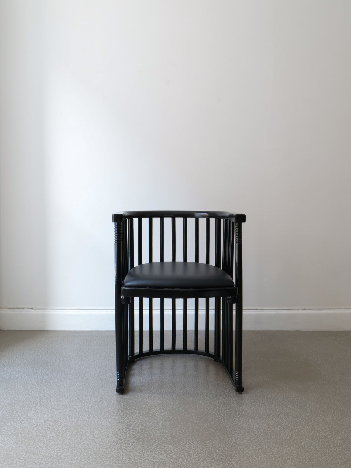 A Bentwood Armchair Josef Hoffman by Collection apart, in black with a simple modern design featuring vertical slats and a curved backrest reminiscent of classic Hoffmann armchairs, sits on a gray floor against a plain white wall. The lacquered bentwood frame adds an elegant touch.