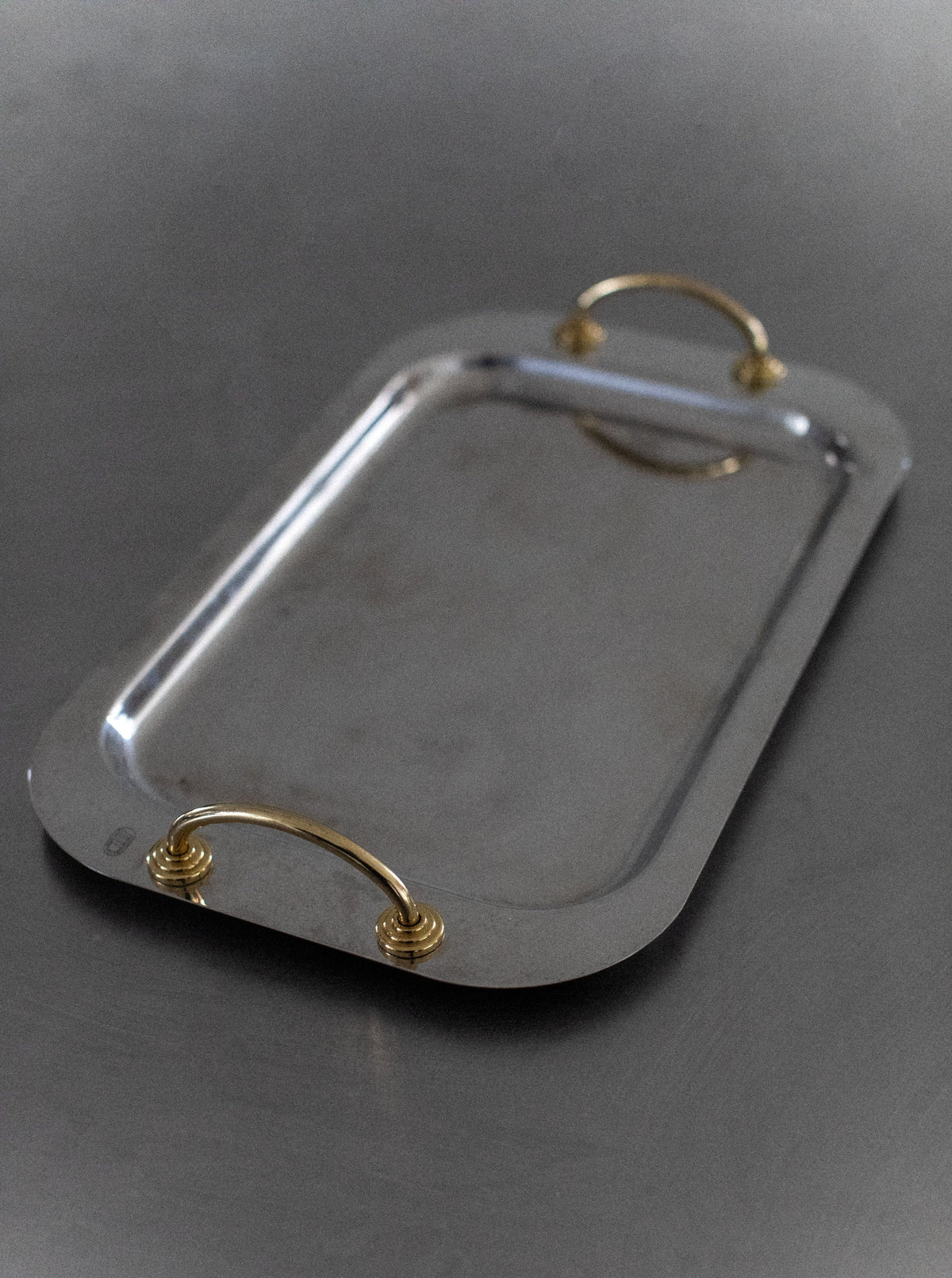 A rectangular Out For Lunch Stainless Steel Tray with rounded corners, featuring two gold-colored handles on either side. The tray rests on a grey surface, reflecting light faintly on its polished finish, showcasing its modern elegance and smooth, contemporary style.