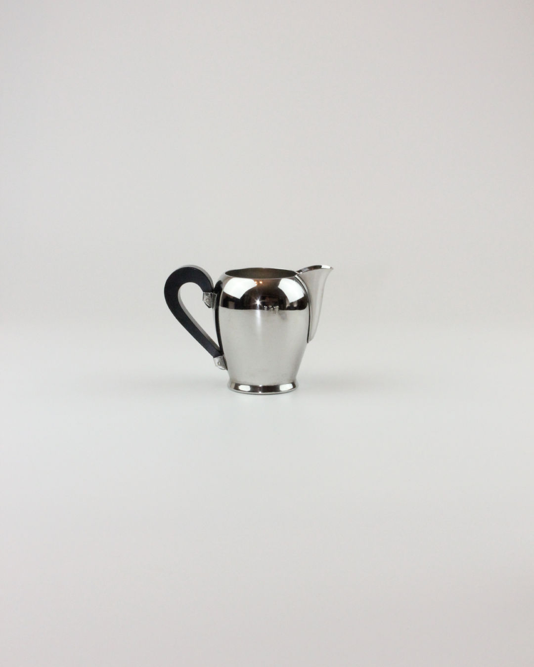 A small, polished silver creamer with a black handle is centered against a plain, light gray background. Showcasing mid-20th-century craftsmanship, the sleek, modern design features a spout on one side and a curved handle on the opposite. This piece hails from the "Bombè" Alessi Tea/Coffee Set by Boga Avante Shop.