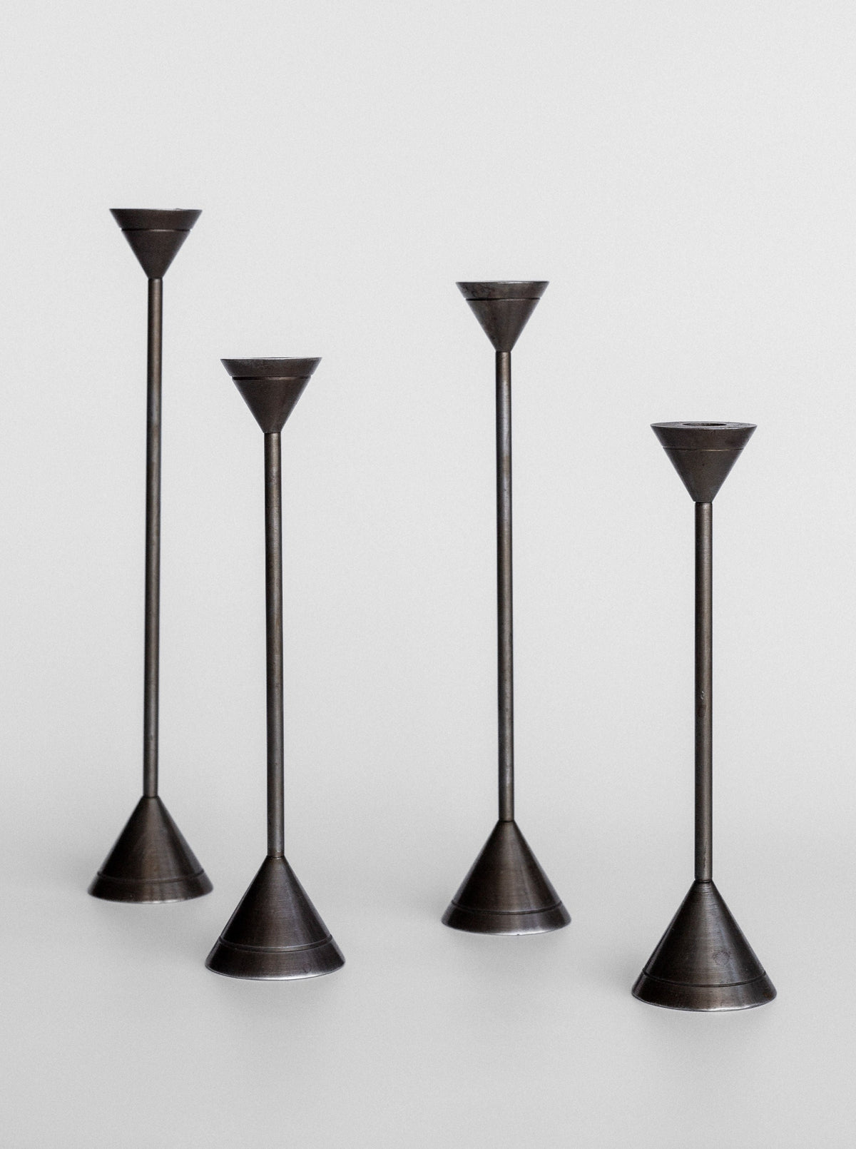 The Spigel Set of Four Postmodern Candle Holders are showcased against a light grey background. Each candle holder features a minimalist, solid metal construction with varying heights, conical bases, and slender stems leading to small, funnel-shaped tops designed to hold candles. Their sleek design exudes postmodern aesthetics.