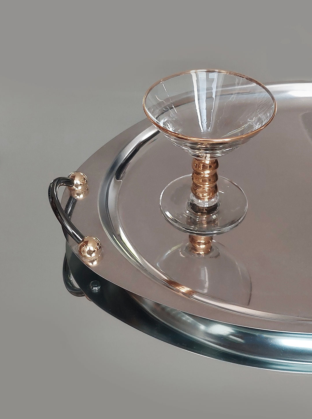 A minimalist image features the sleek, modern Medium Oval Steel Tray by Les Objoies with a glossy surface. Resting on the tray is an elegant cocktail glass with a gold-toned stem. The decorative serving tray's handle is adorned with a small, gold sphere. The background is plain grey.