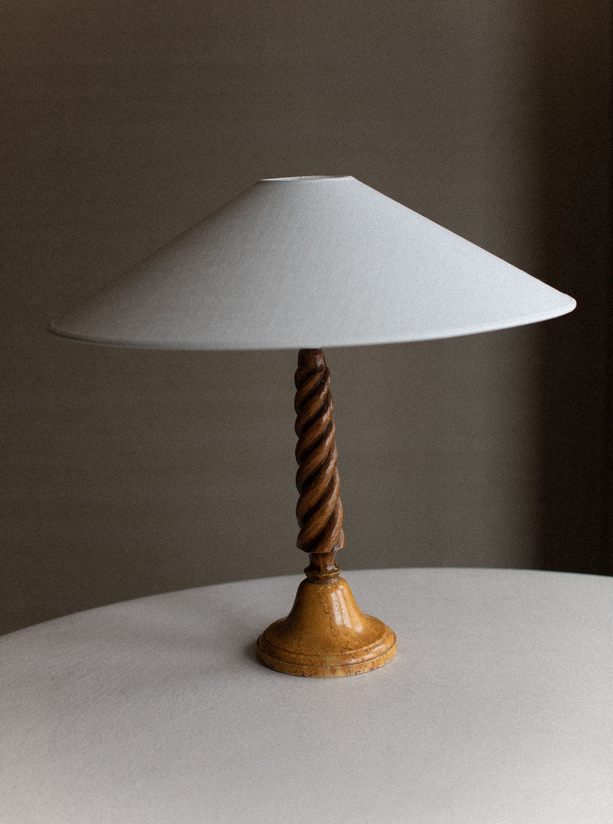 A Twisted Wooden Lamp by Out For Lunch with a white conical shade and a twisted wooden base sits on a plain white surface. The background is dark and out of focus, emphasizing the artisanal craftsmanship of the lamp's design.