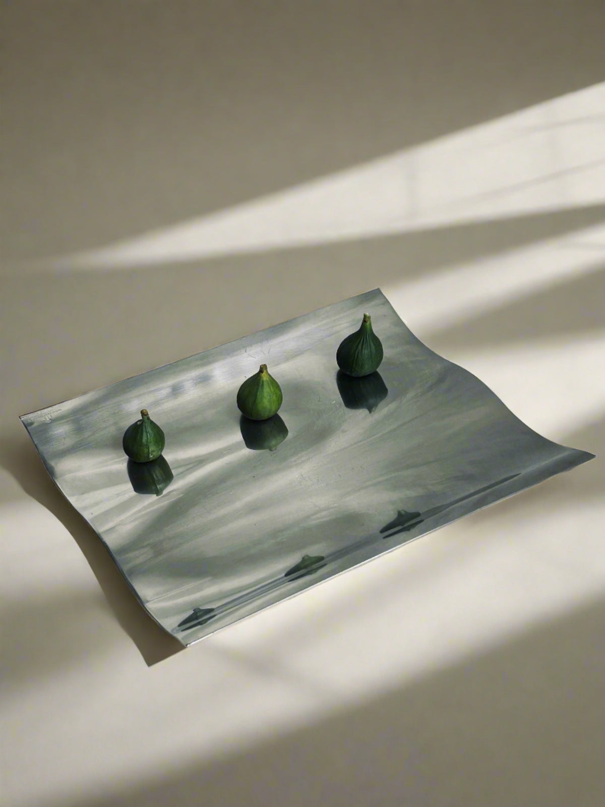 The elegant Wave Tray by Royal Copenhagen from Flock Studio, crafted in stainless steel, holds four small green figs and casts subtle shadows on a neutral-toned surface. Soft light and shadows form an elegant, minimalist composition with a modern twist.
