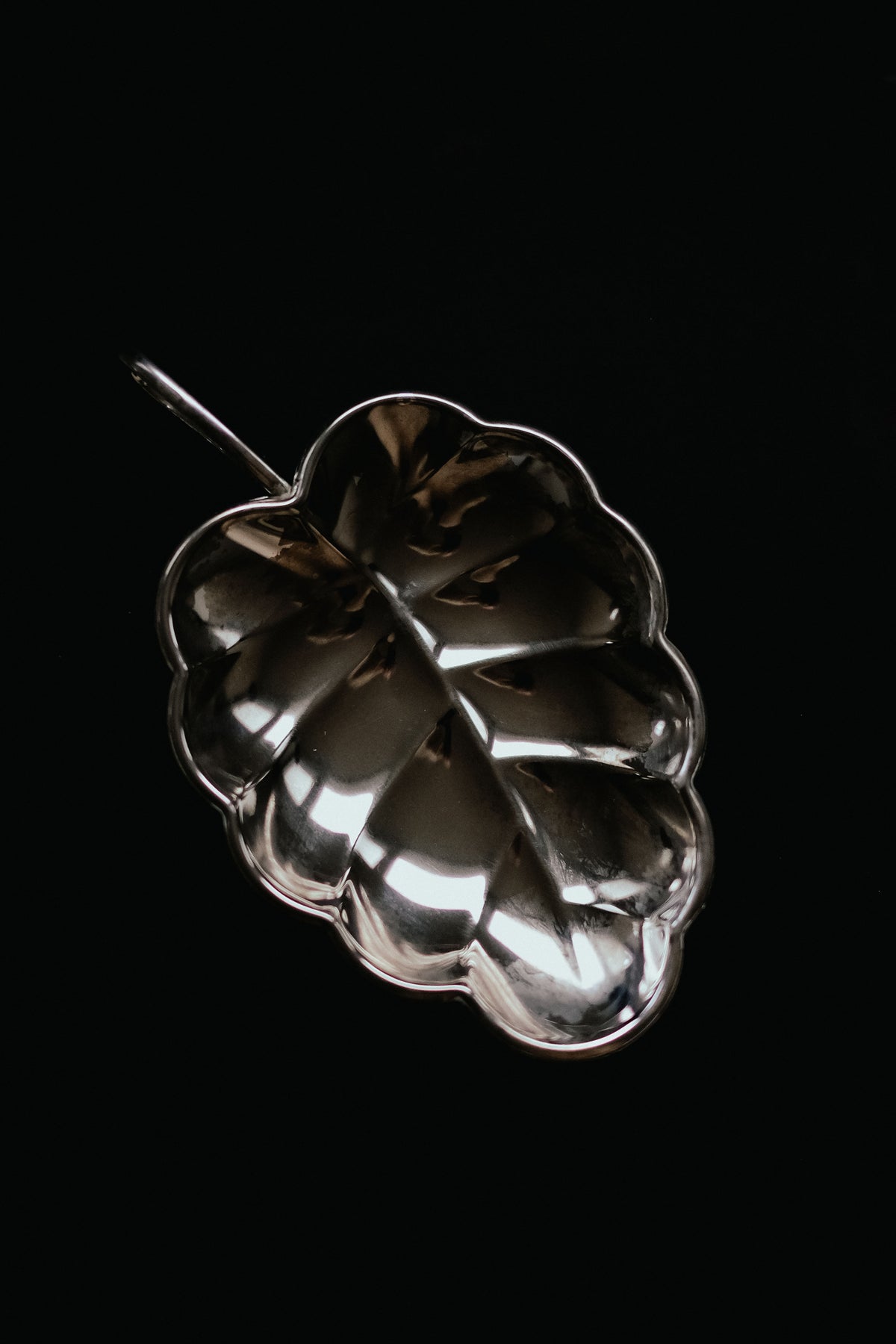 The Serving Dish Leaf from septembre studios features a shiny silver, leaf-shaped design with a stem-like handle and distinct vein patterns, set against a black background for a vintage appeal.