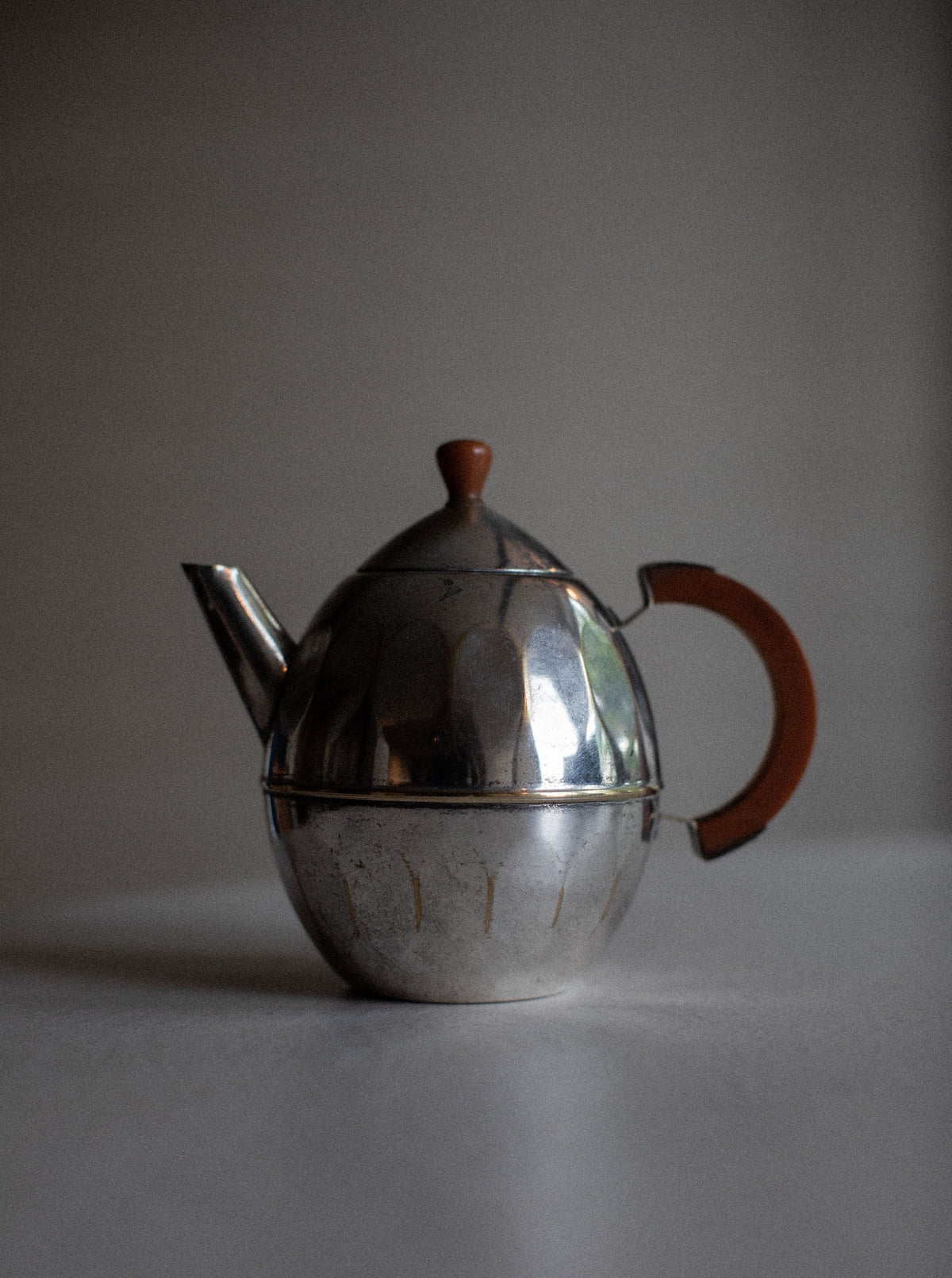 A vintage **Out For Lunch Small Teapot** with a round body and a slightly conical lid. It has a short spout and a curved handle with a brown wooden grip. The surface of the teapot is slightly reflective and shows visible wear, adding to its timeless elegance. It is set against a plain, muted background.