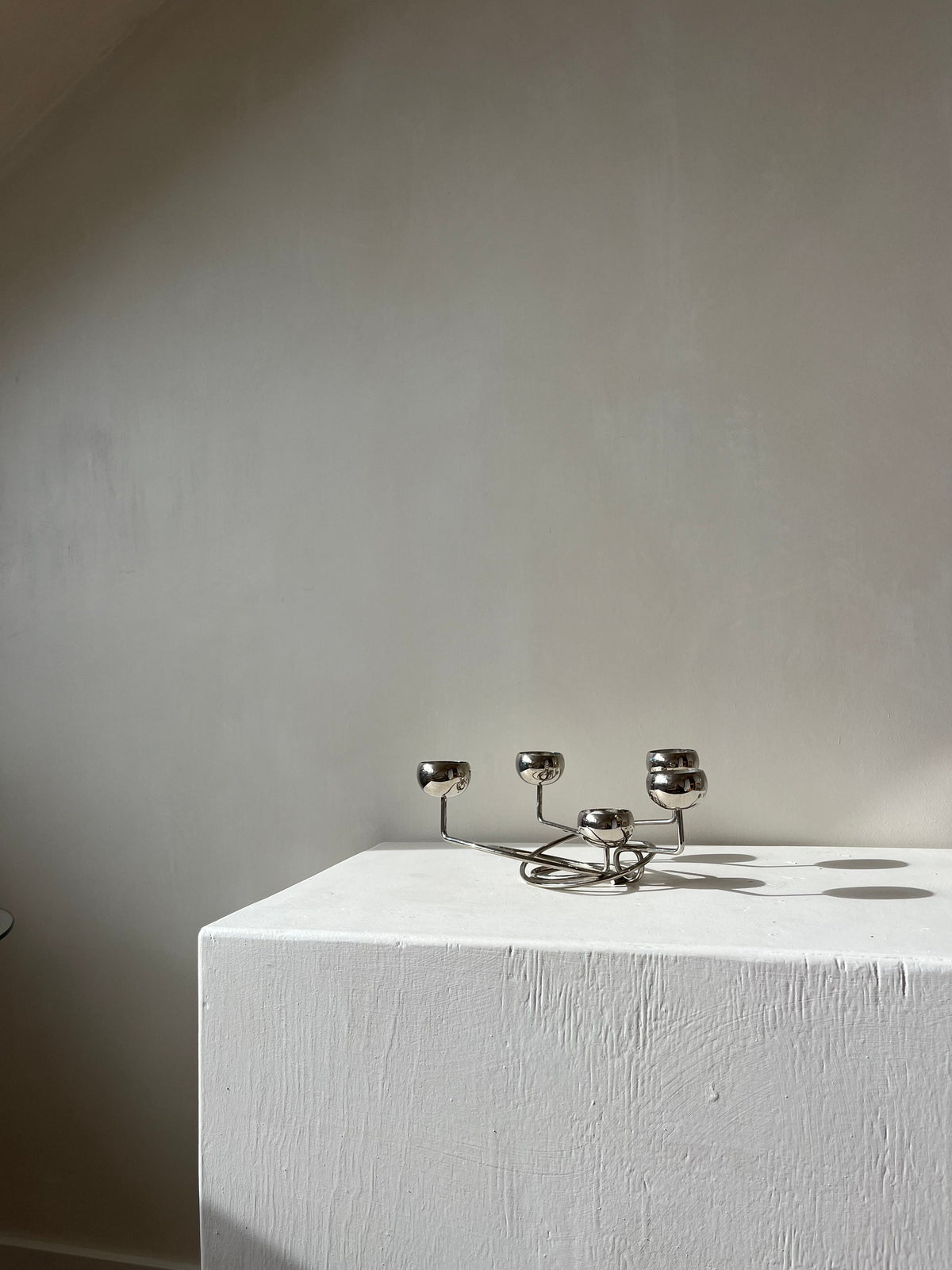 A Chrome Candleholder by septembre studios rests on a white plinth in a room with soft, neutral-colored walls. Sunlight casts gentle shadows at unique angles, creating a calm and serene atmosphere.