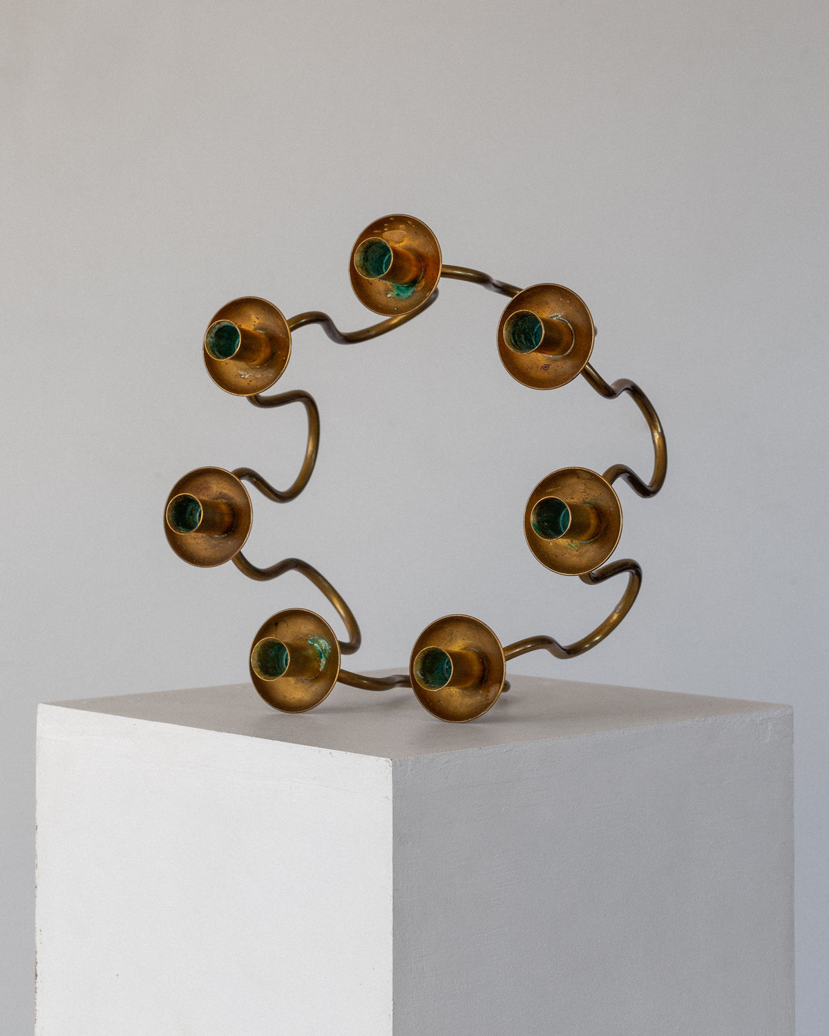 The Scandinavian Brass Candleholder by Spigel, reminiscent of Lars Holmström's modern sculptures, features wavy bronze rods with golden spheres and green centers. It exudes a vintage allure atop a white pedestal against a plain light gray background.
