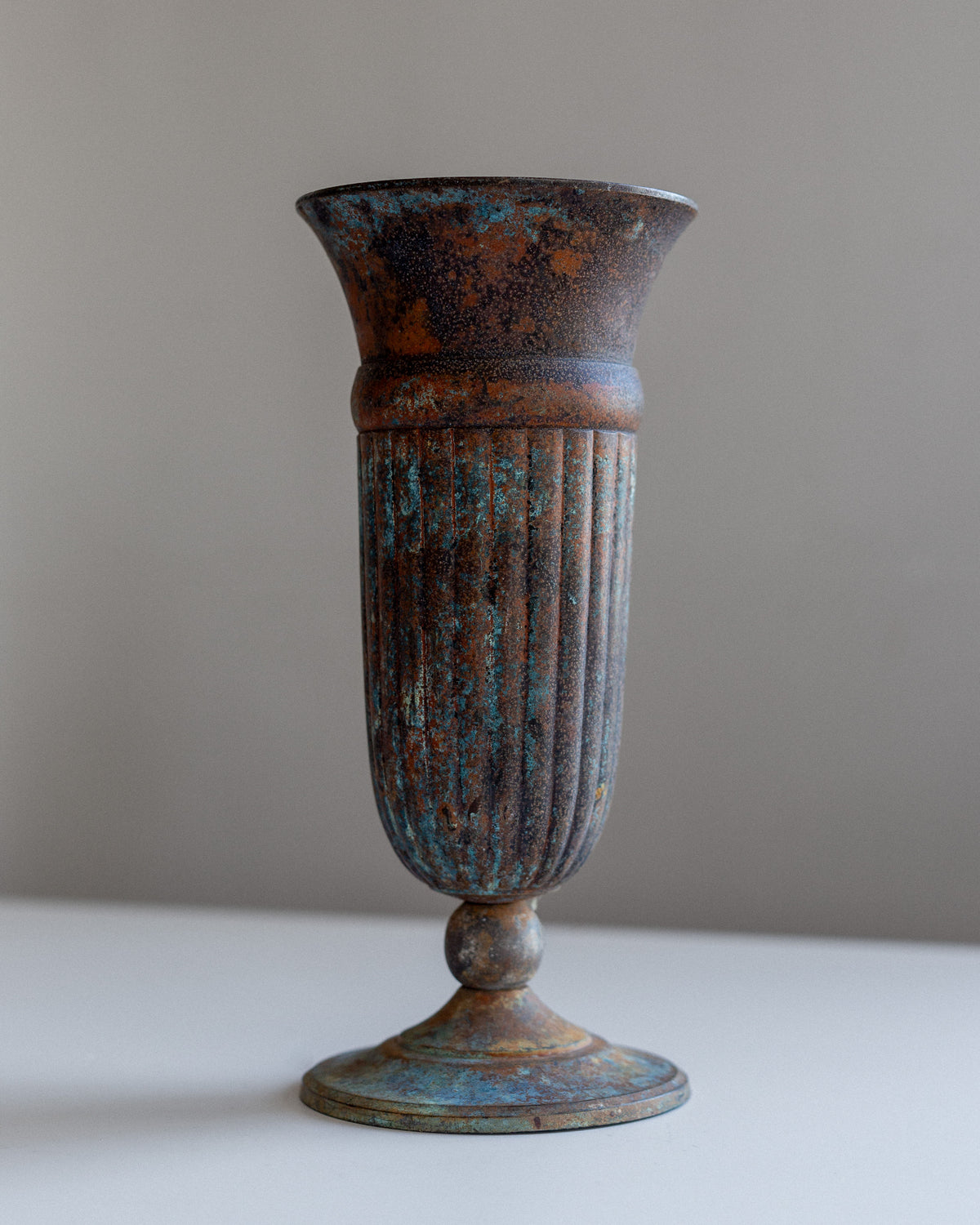 A tall, Spigel Vintage Cast Iron Vase with a fluted design stands on a plain surface against a gray background. The vase showcases a weathered patina, antique character, and a rounded base.