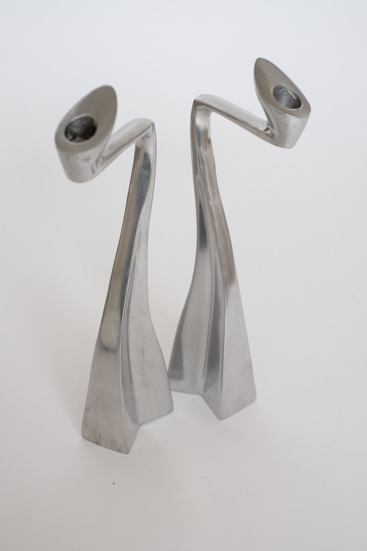 The Pair of XL Candle Holders by Matthew Hilton from septembre studios, featuring a modern abstract design in silver with elegant curves, serve as stunning collector's pieces against a neutral background.