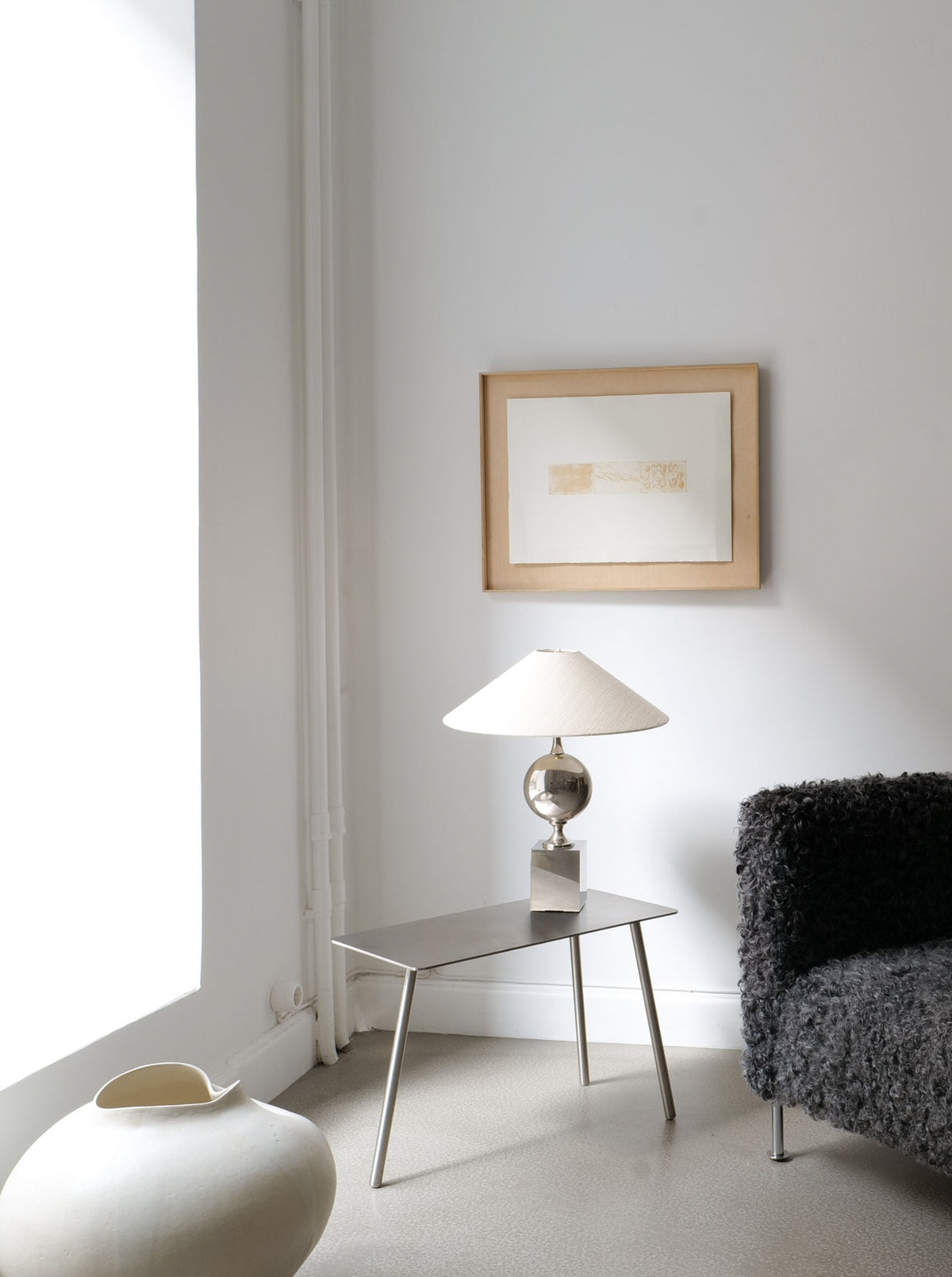 A minimalist room features a grey, textured armchair with metallic legs, an angular Stick Table Jesper Morrison 2000 by Collection apart with a white lamp, and framed abstract artwork on a white wall. A large, round, white ceramic vase sits on the floor near a tall, sunlit window.