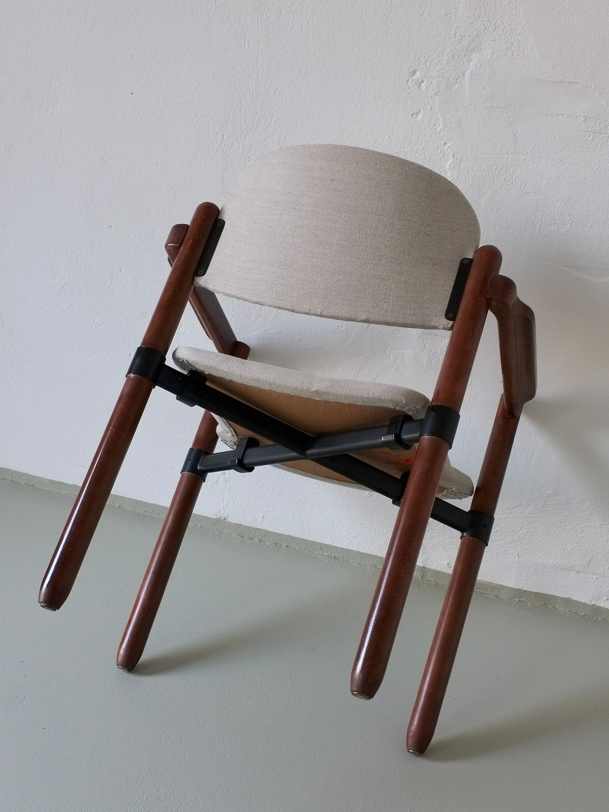 A Fröscher Sitform armchair by Veter Vintage, featuring a wooden frame and light beige cushioned seat and backrest typical of 1970s design, is upside down against a textured white wall. It's supported by black metal brackets seen on stacking armchairs from the 1970s Germany set.
