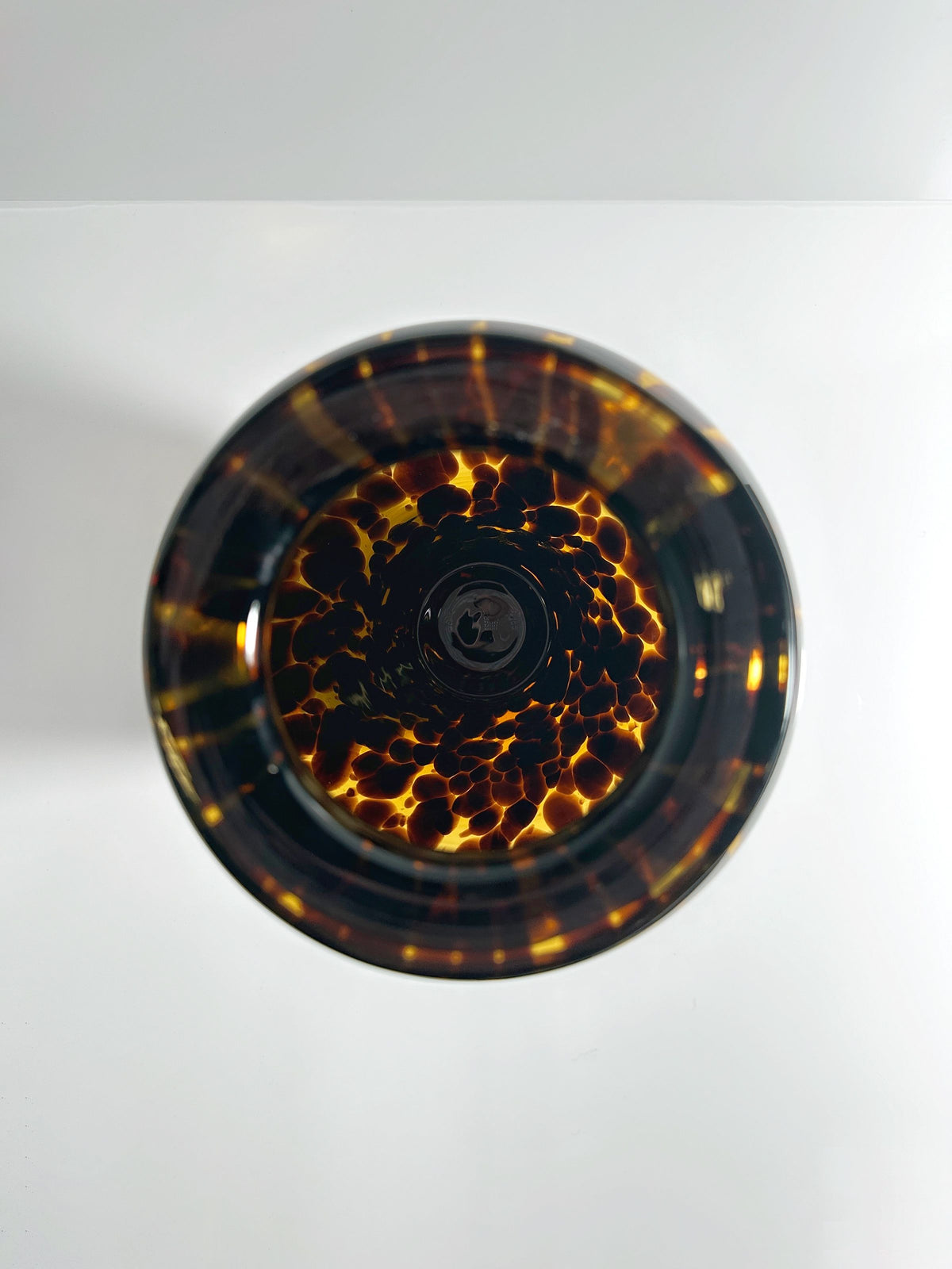 The top view of a glass filled with a dark liquid, its surface patterned with light reflections and bubbles resembling amber, mirrors the intricate design of Dodo Vintage's Tall Murano Tortoise Vase against a smooth white background.