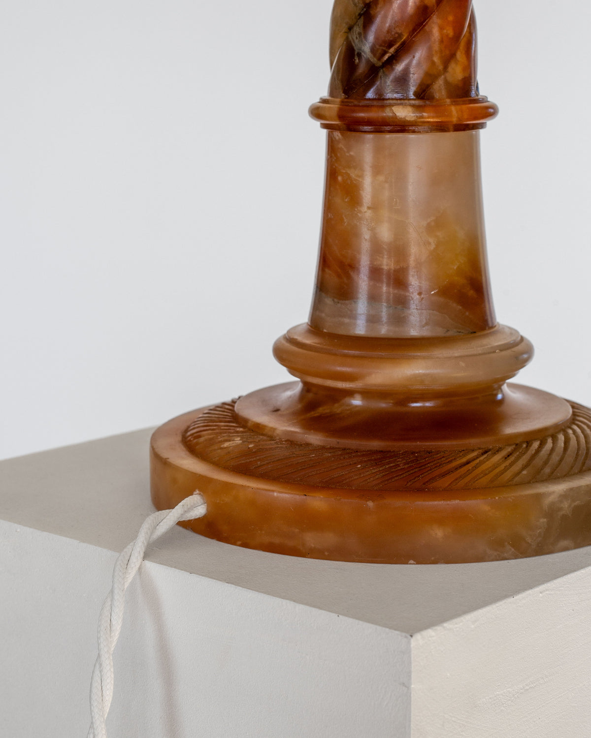 A close-up photograph features an exquisitely hand-carved base of the "Antique Alabaster Table Lamp, C. 1900" by Spigel, showcasing its stunning shades of brown and amber. The lamp rests on a white pedestal with a white cord emerging from the base, casually draped across the pedestal's surface, highlighting its classic and timeless aesthetic.