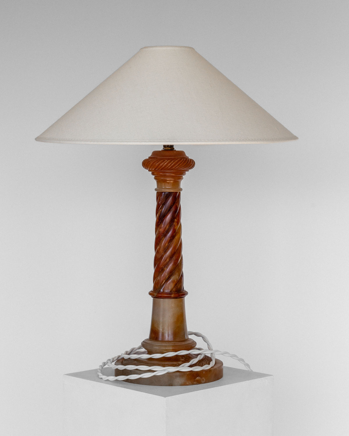 The Spigel "Antique Alabaster Table Lamp, C. 1900" features a beige cone-shaped lampshade and a decorative, twisting wooden base. It stands on a white pedestal, exuding a classic and timeless aesthetic. An electrical cord is loosely coiled around the bottom of the lamp.