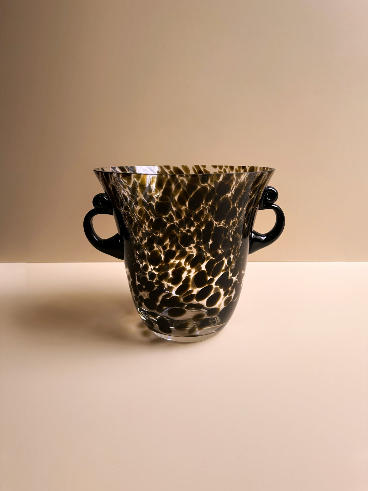 The Art Deco Champagne Bucket / Vase Tortoise Glass by Dodo Vintage is a handcrafted Italian piece, featuring an ornamental design with a flared rim and two small, curved handles on each side. The tortoise glass vase showcases a translucent surface adorned with intricate marbled patterns in dark brown and black hues. It is displayed against a plain, beige background.