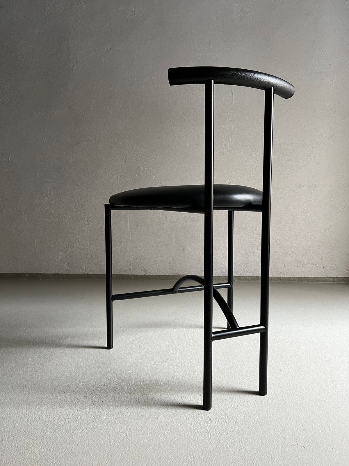 A minimalist modern black stool with a sleek design, featuring a thin, curved backrest and metal legs. The faux leather upholstery adds a touch of sophistication. Positioned in front of a plain, light gray wall on a smooth, light-colored floor, no other objects are visible in the scene. 4 Postmodern "Tokyo" Chairs by Rodney Kinsman | Italy | 1980s by Veter Vintage provide an elegant yet contemporary seating solution.